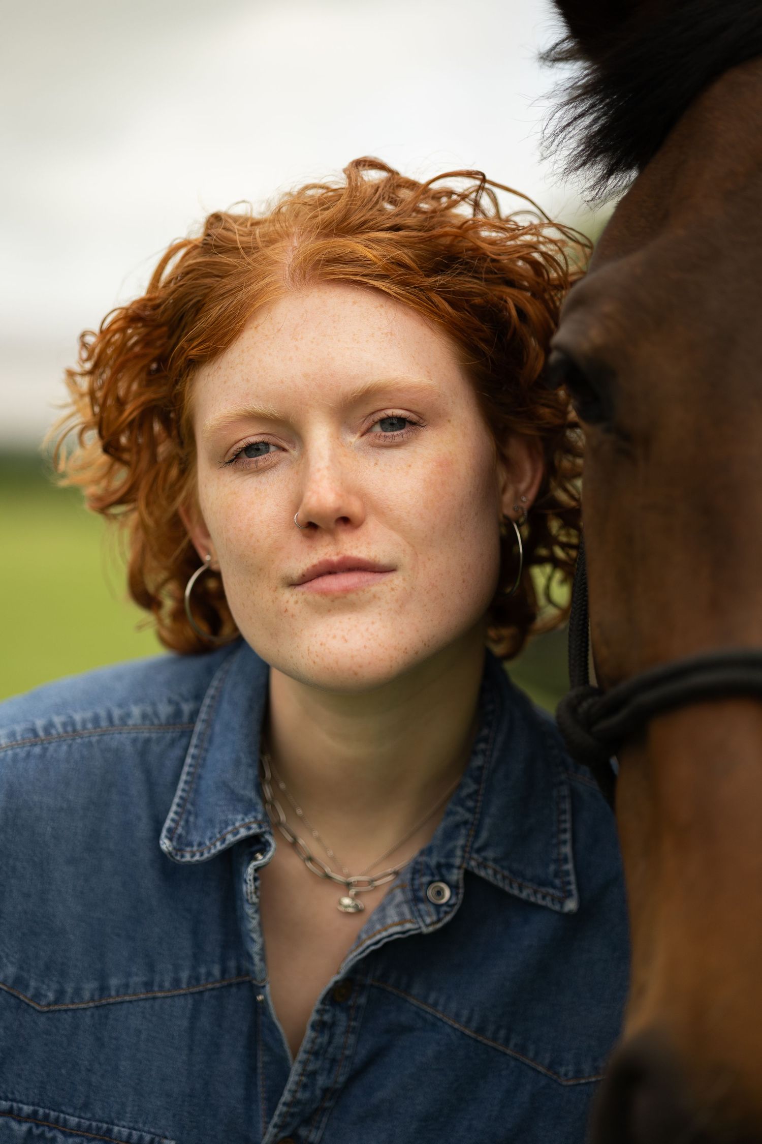 Close-up portrait of a curly red-haired woman with freckles leaning against a brown horse outdoors.