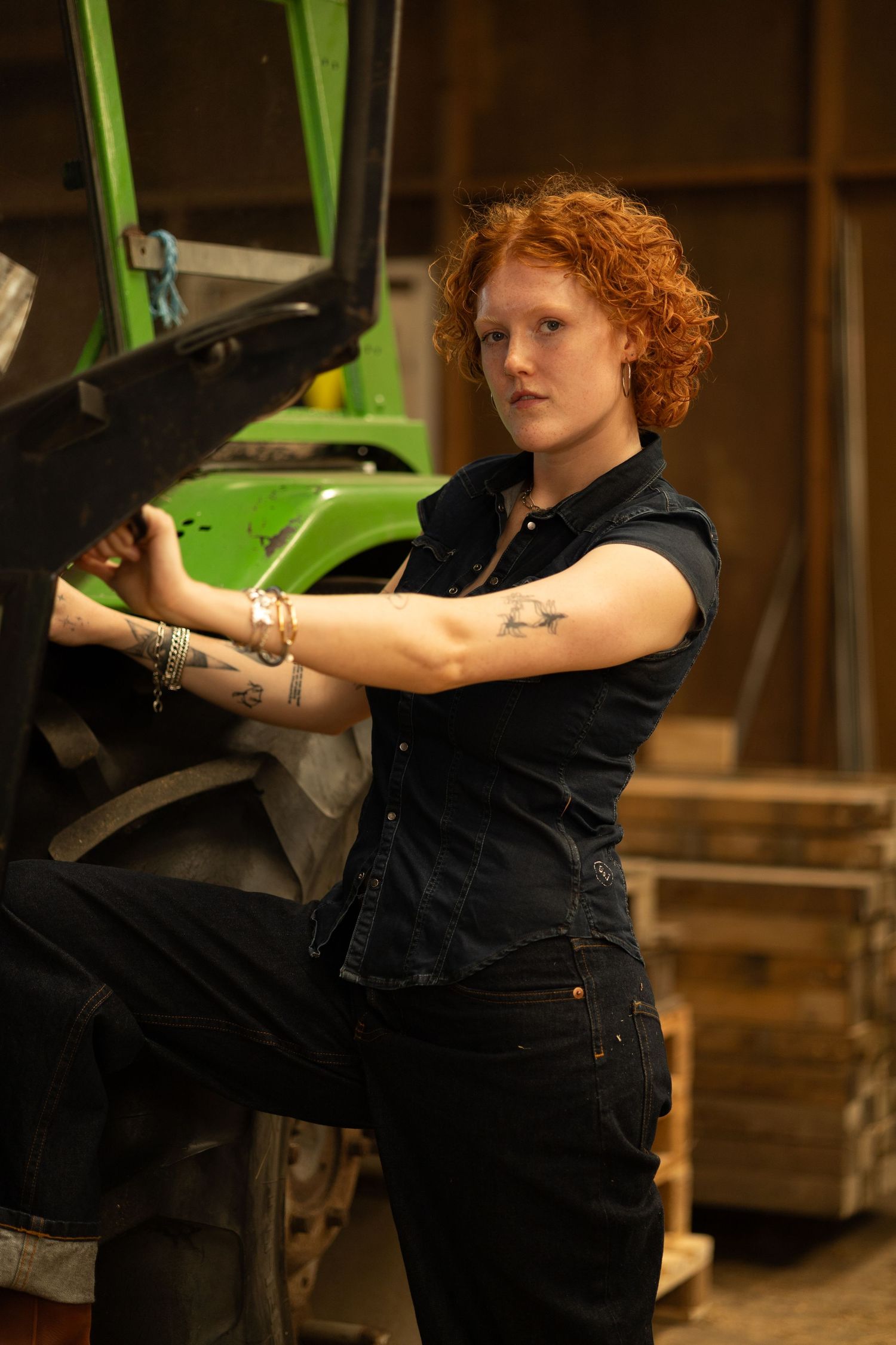Red-haired woman in black denim overalls working on a green tractor in a barn, showing tattoos on her arms.