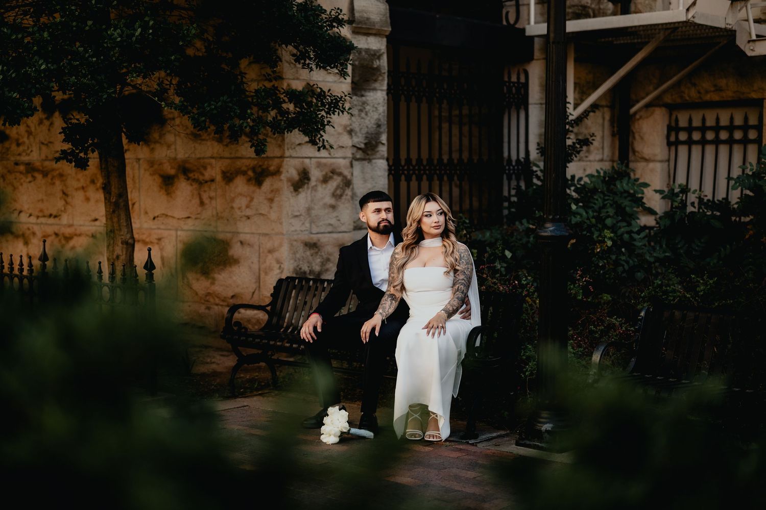 Wedding couple sitting together on bench surrounded by lush greenery against a warmly lit stone wall.