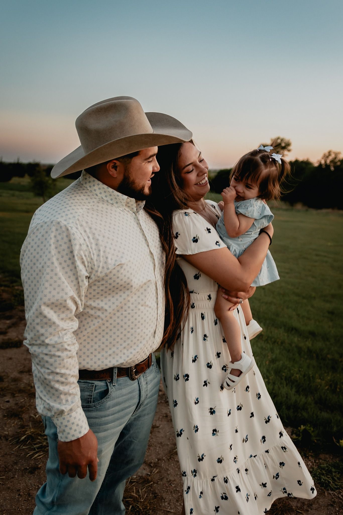 Family of three with father in cowboy hat holding toddler daughter at golden hour outdoors.