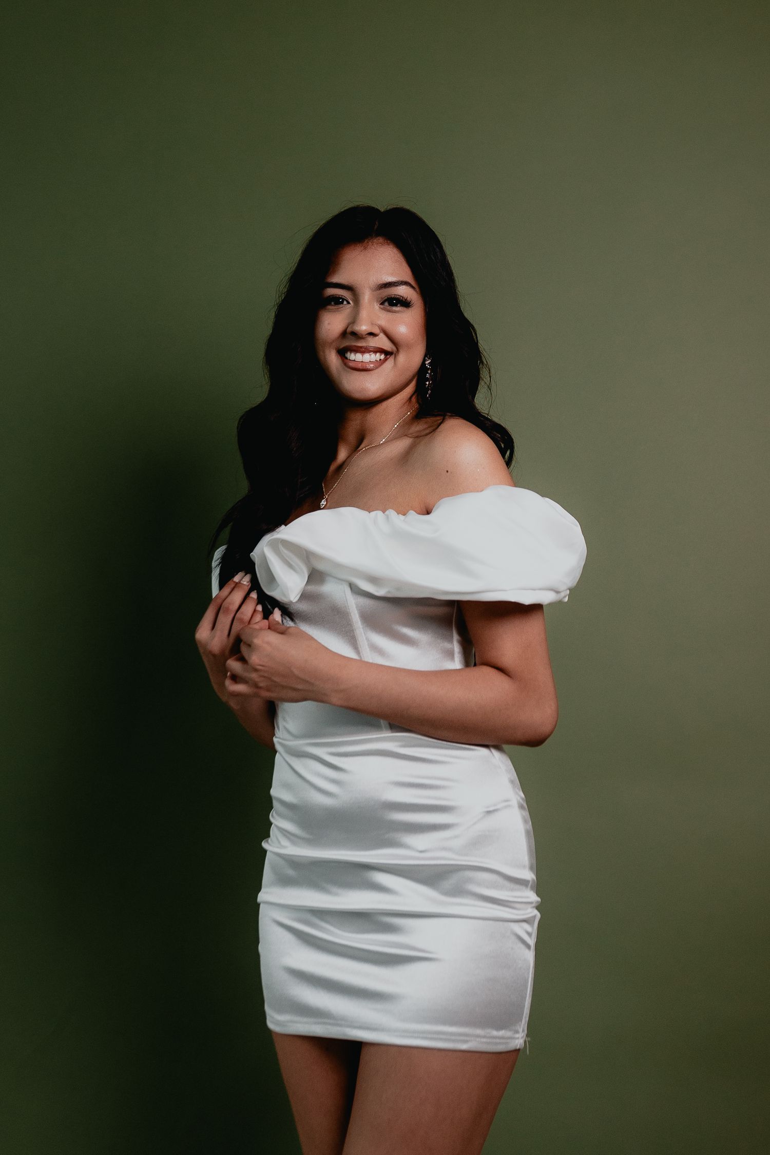 Smiling woman in white off-shoulder satin dress posing against olive green background.