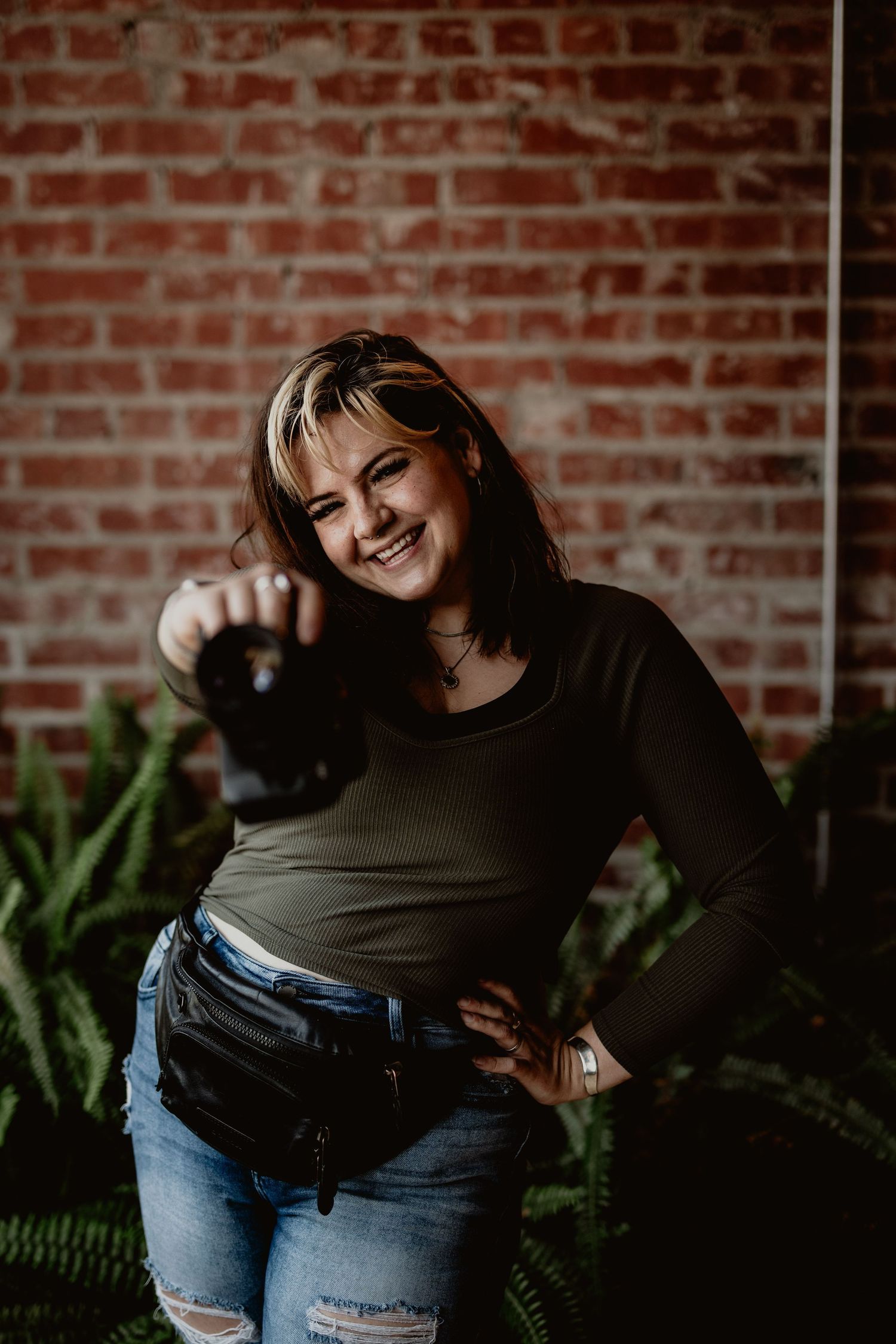 Smiling woman in olive top and jeans holding a camera toward the lens near a brick wall.