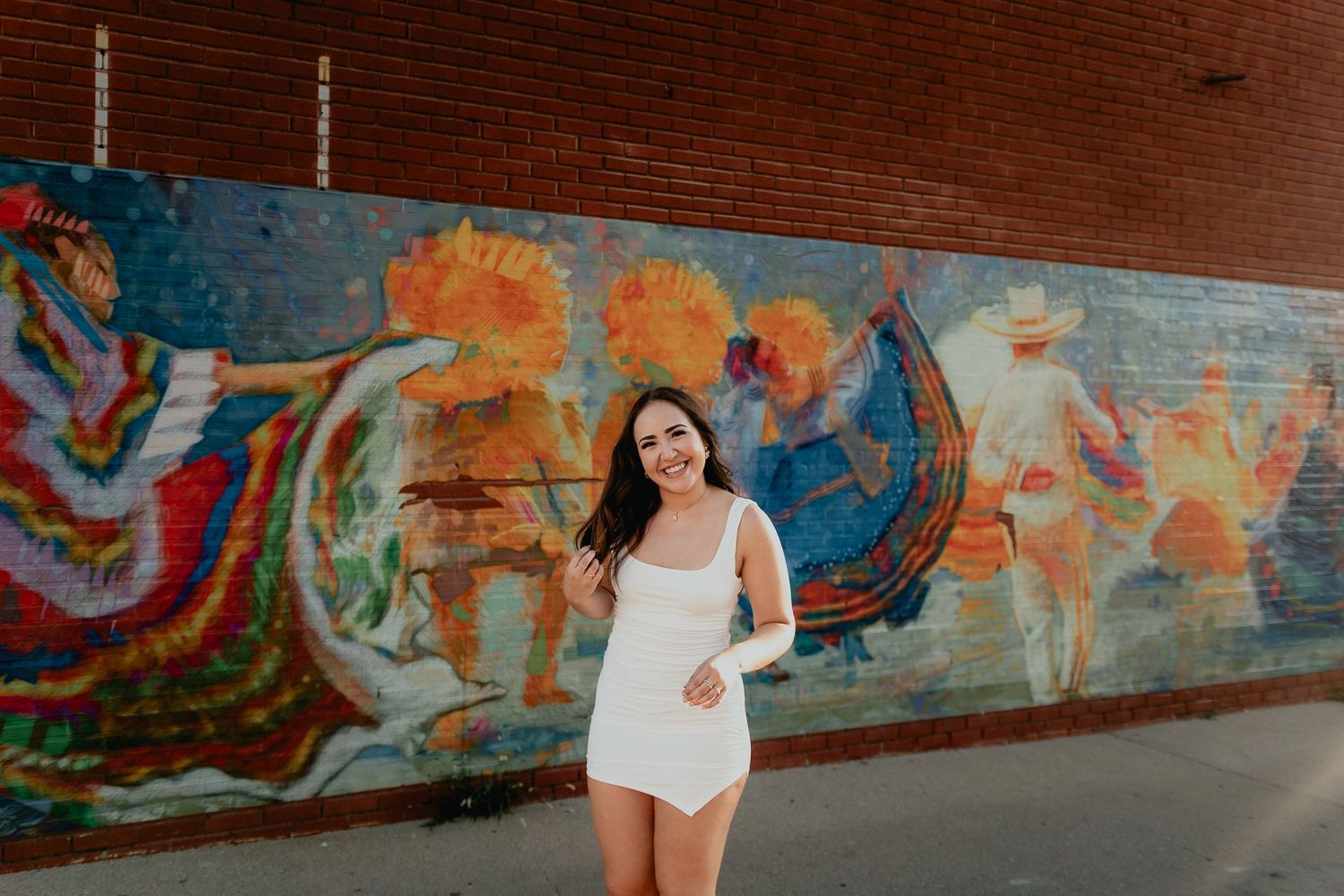 Smiling young woman in white romper poses in front of vibrant colorful street mural on brick wall.