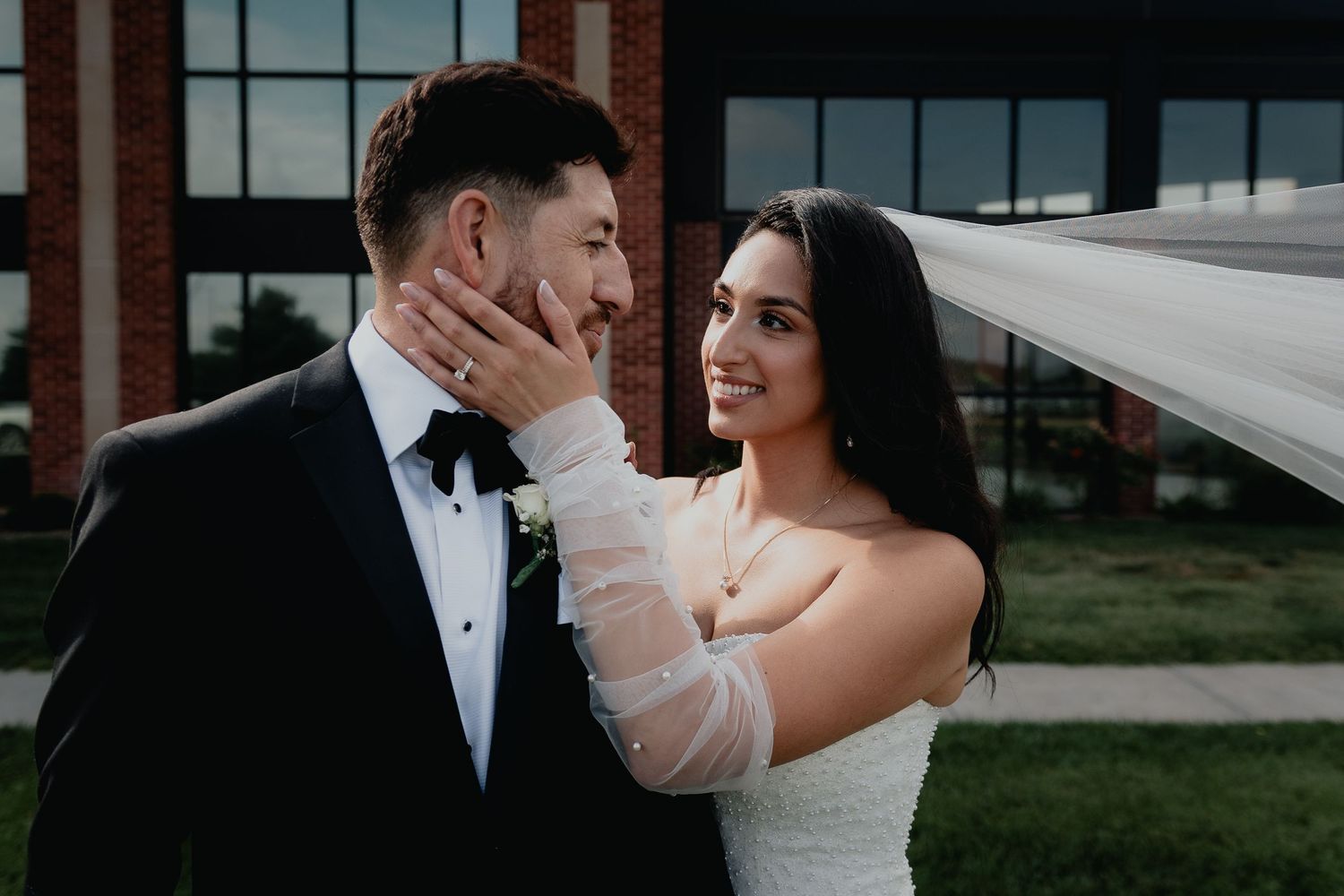 Elegant bride in white dress tenderly cups smiling grooms face during outdoor wedding portrait session.