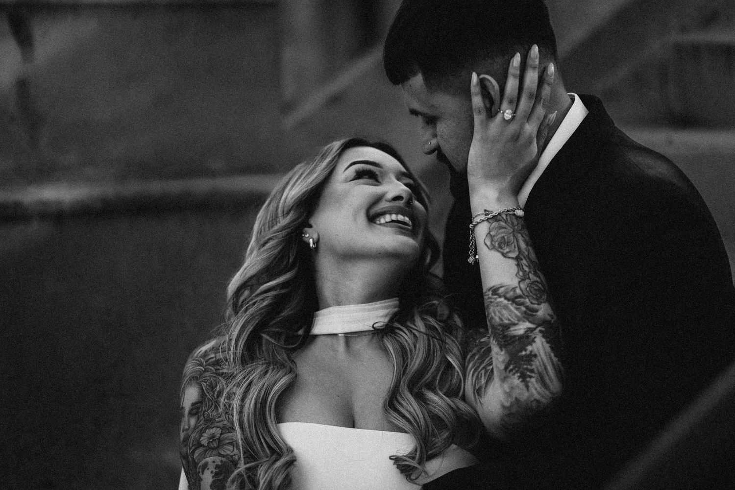 Black and white intimate wedding portrait of tattooed bride laughing with groom in formal suit.
