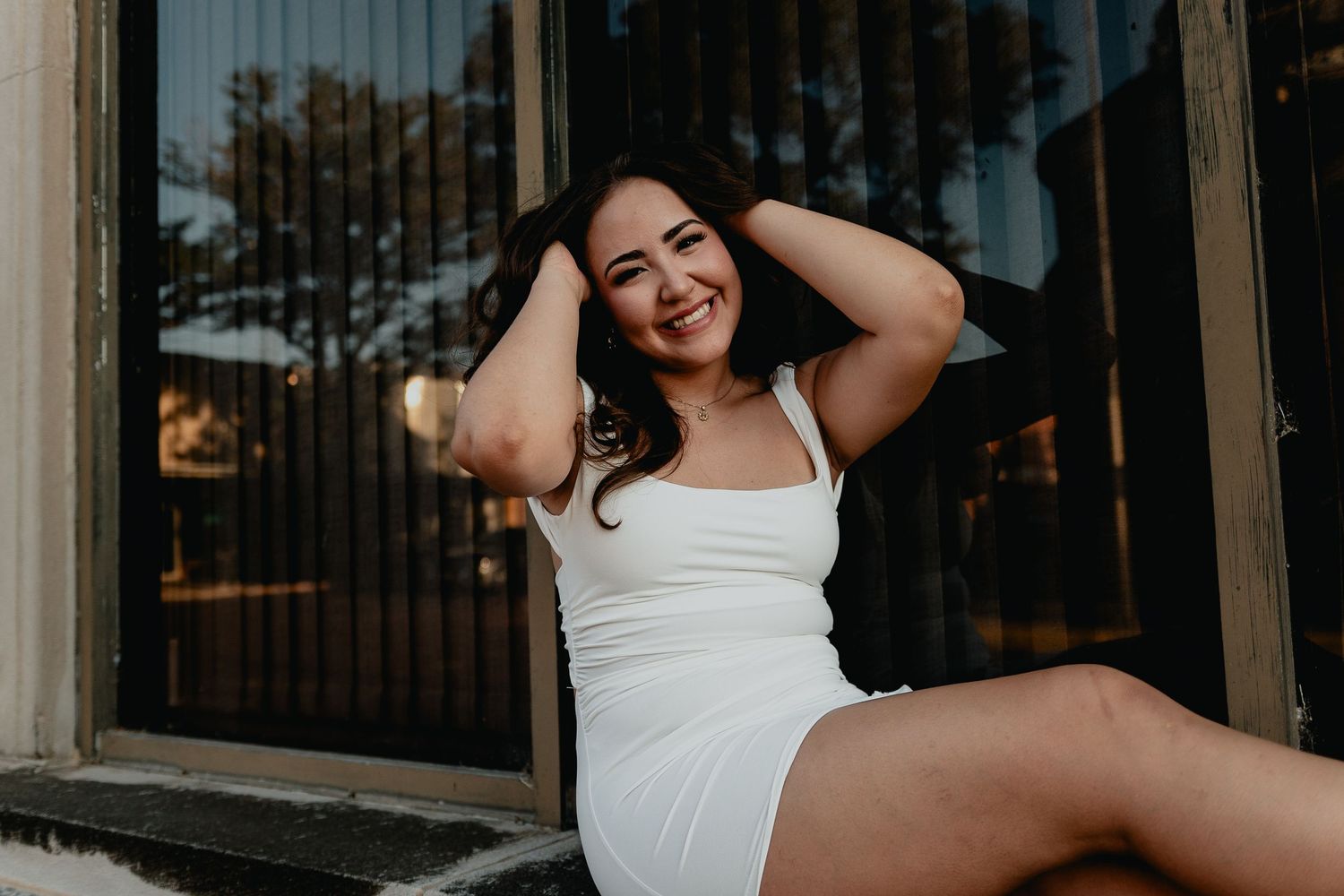 Happy young woman in white mini dress sits smiling with hands behind head near sunlit storefront.