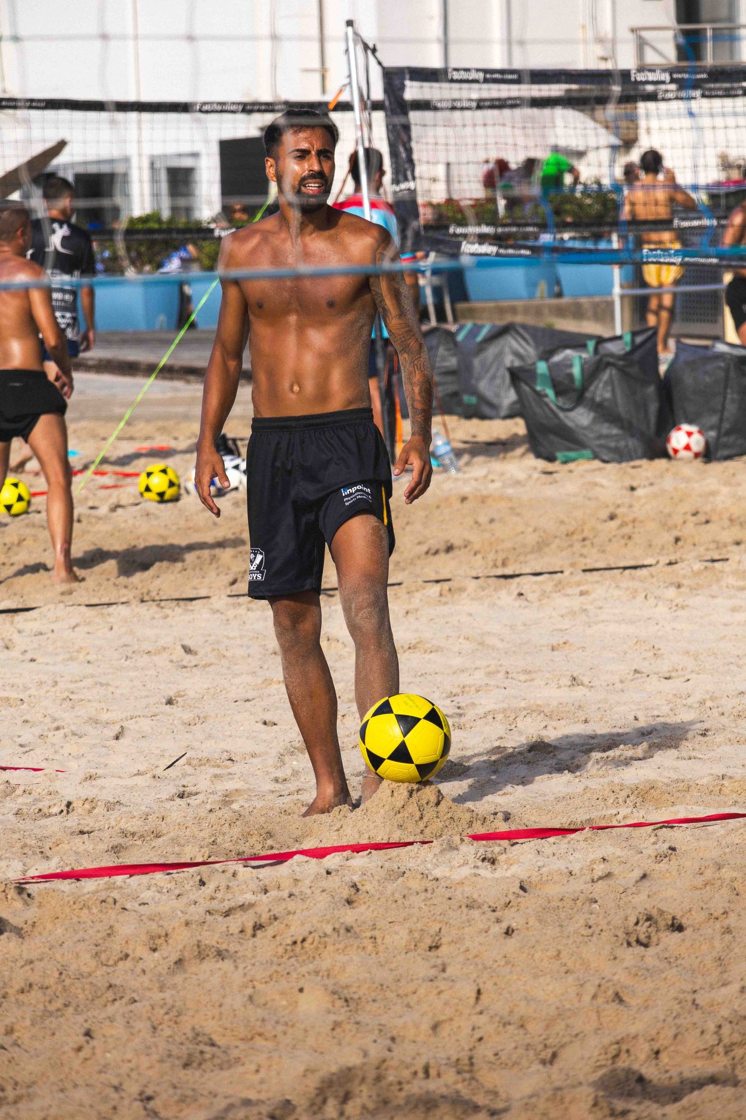 Athletic player with soccer ball on beach volleyball court during sunny day with nets and equipment in background.
