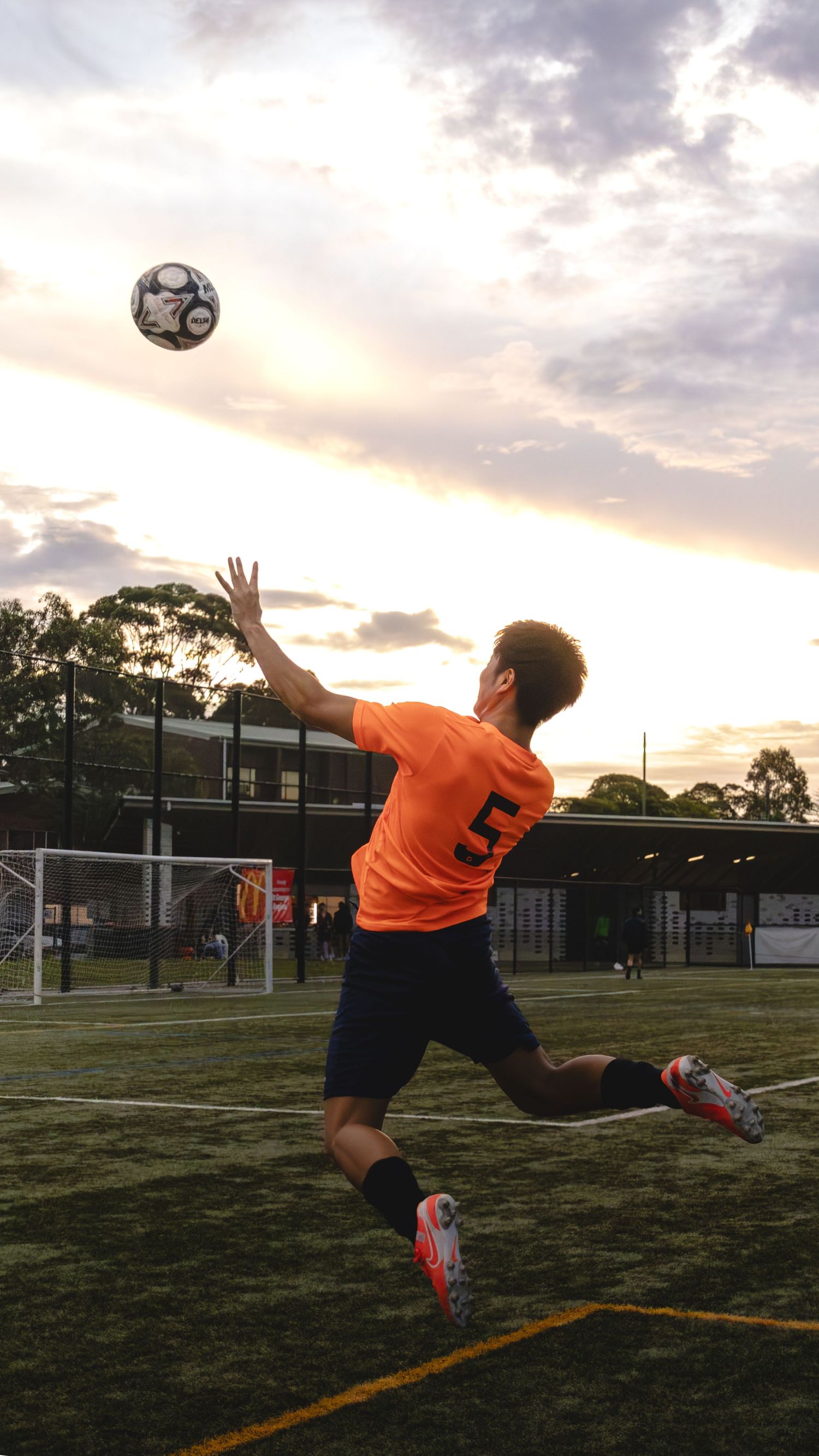 Soccer player in orange jersey performs an acrobatic bicycle kick during sunset on a grass field.