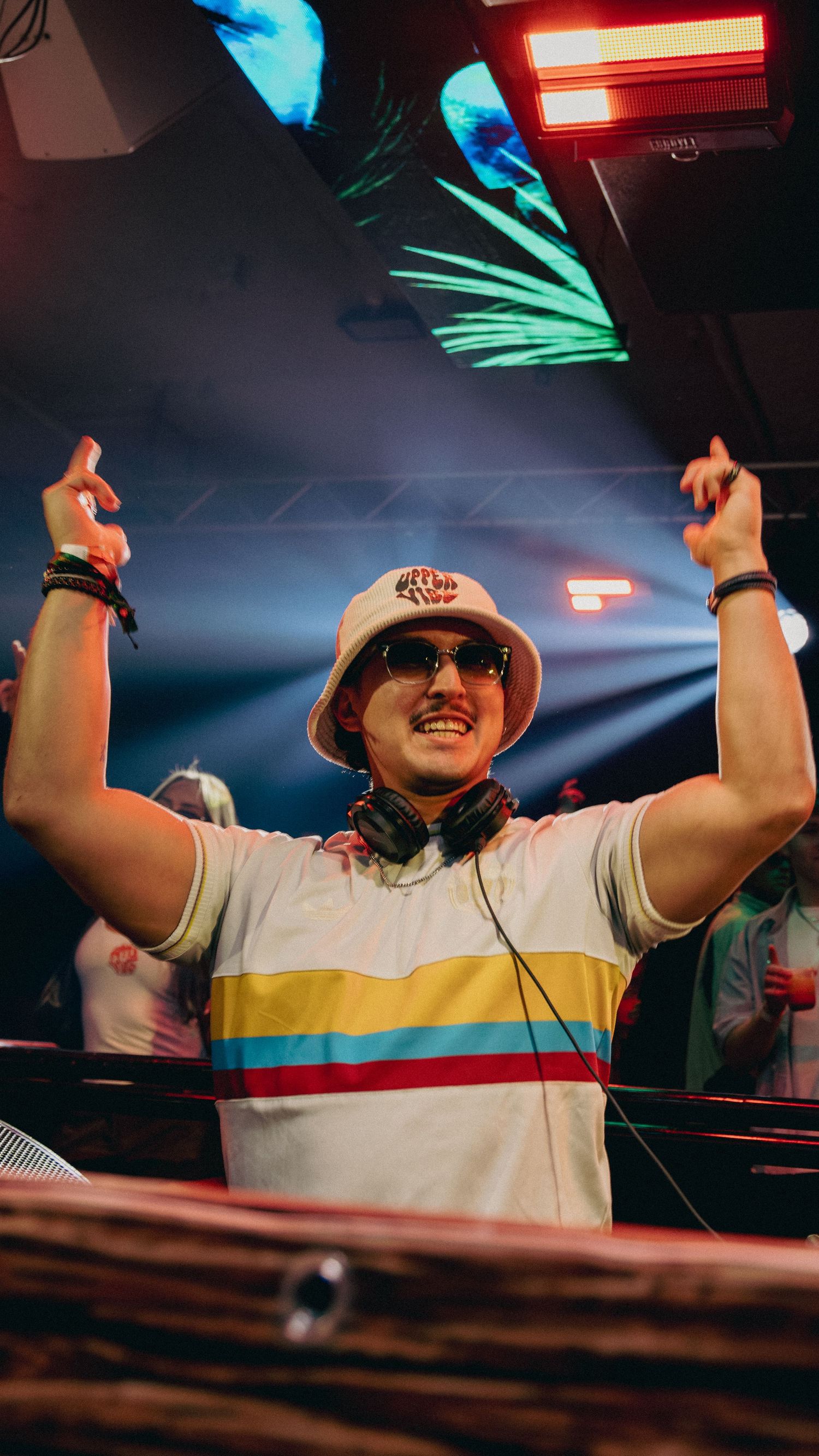 Smiling DJ with headphones around neck, bucket hat, and striped polo raises hands at a vibrant nightclub event.