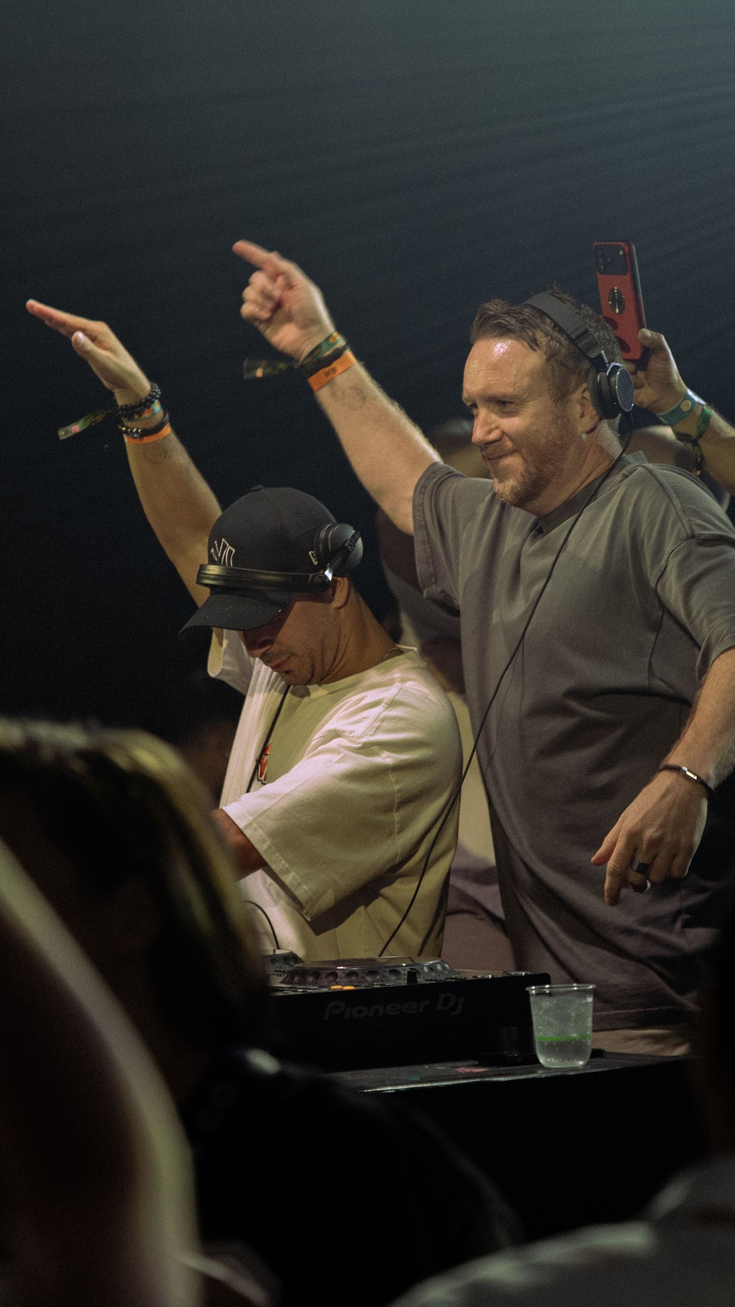 Two DJs performing at a live music event, one raising hand with headphones and Pioneer DJ equipment visible on stage.