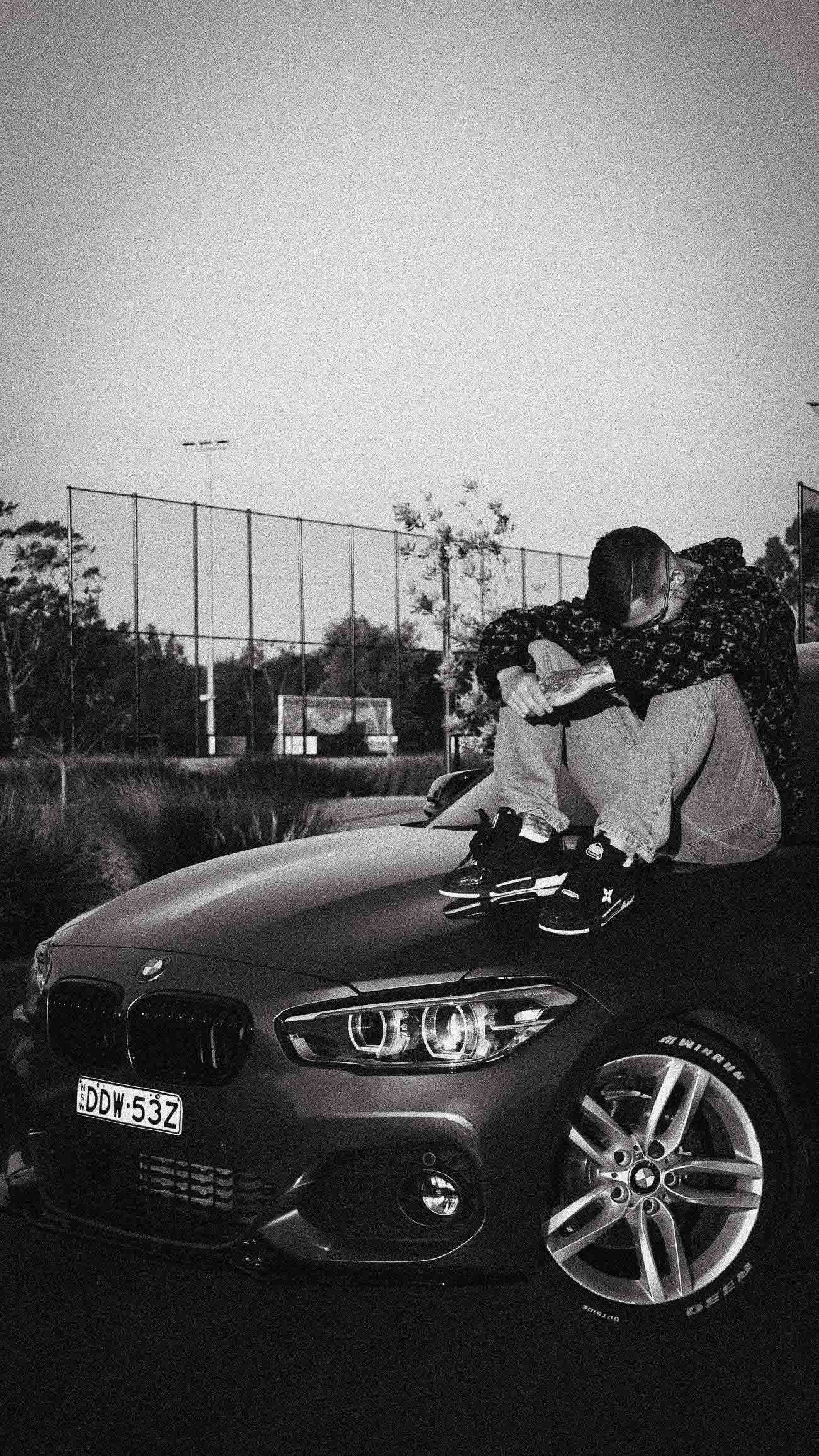 Person sitting on hood of black BMW 1 Series car in black and white photo outdoors near sports field.