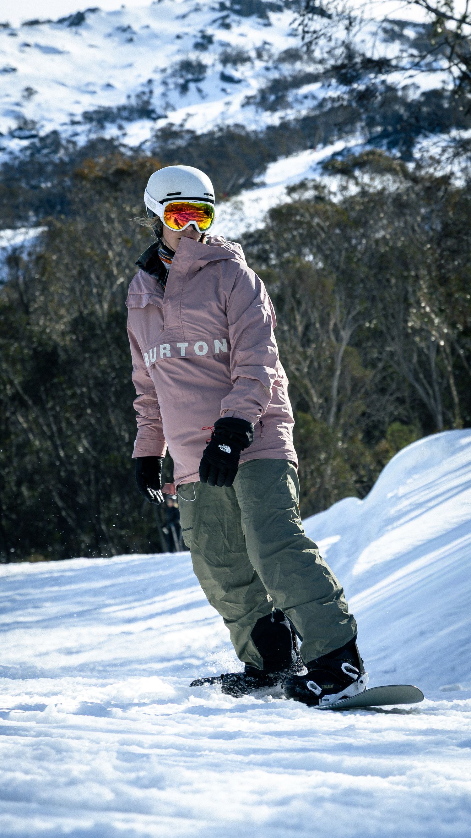 Snowboarder in pink Burton jacket and olive pants riding snowy mountain slope wearing white helmet and orange goggles.