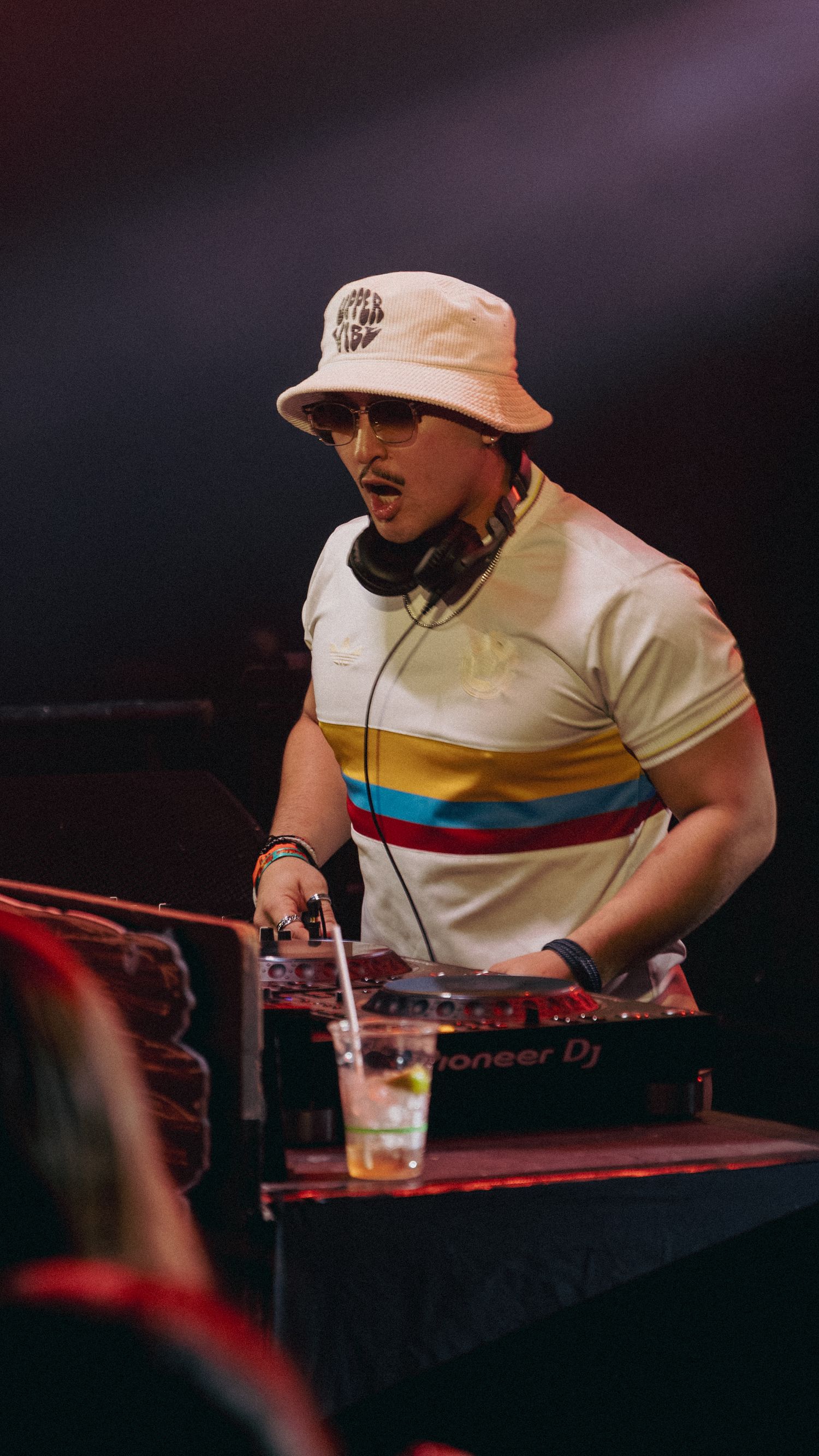 A DJ wearing a bucket hat and striped shirt operates Pioneer DJ equipment on stage under dramatic lighting.