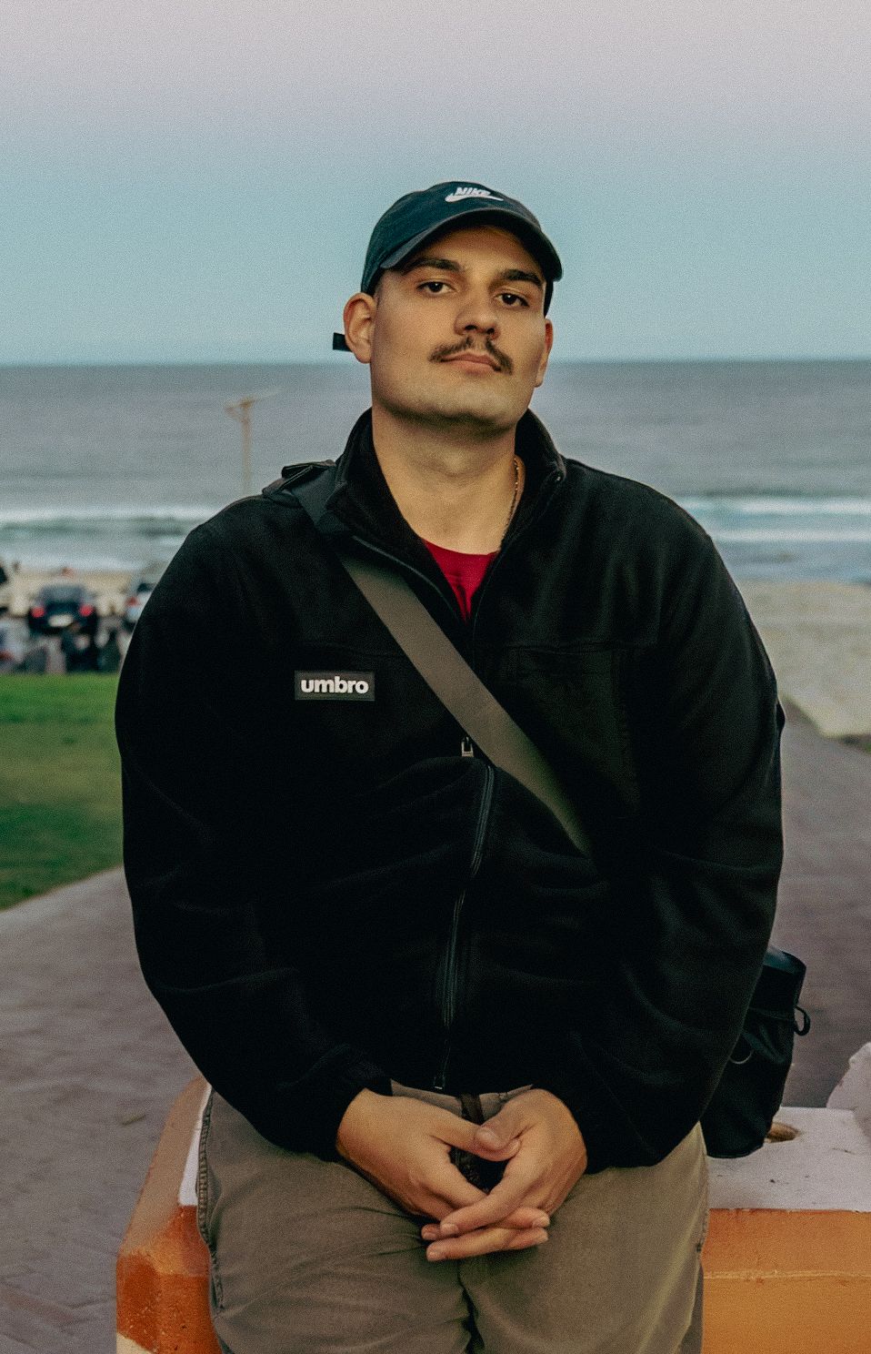 Young man with mustache wearing black Umbro jacket and cap, sitting near ocean beach with calm sea in background.