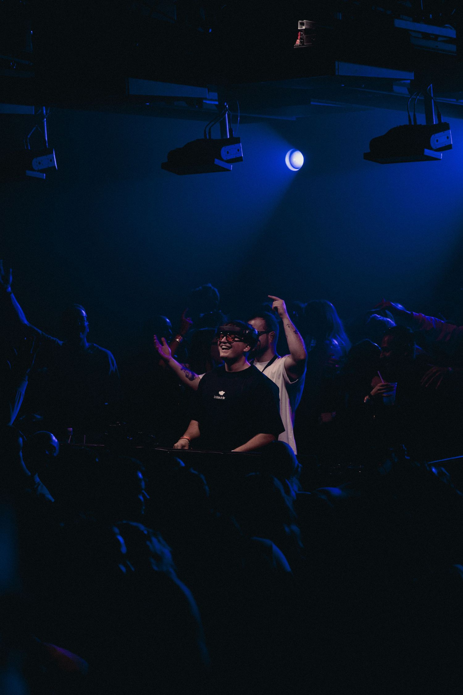 DJ performing at a nightclub with blue stage lighting and crowd dancing with raised hands in the background.