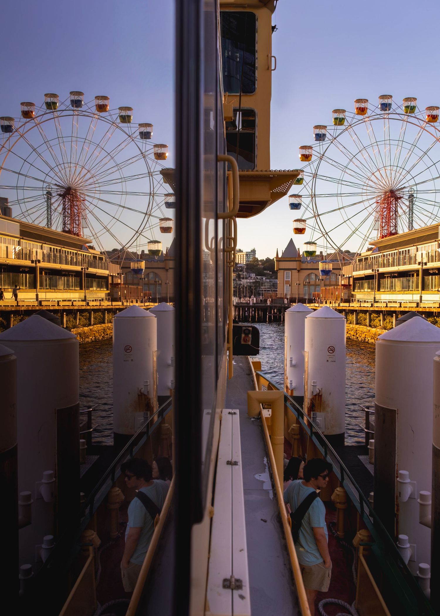 Reflective view of a Ferris wheel at sunset with boats docked along a waterfront pier creating a mirror image.
