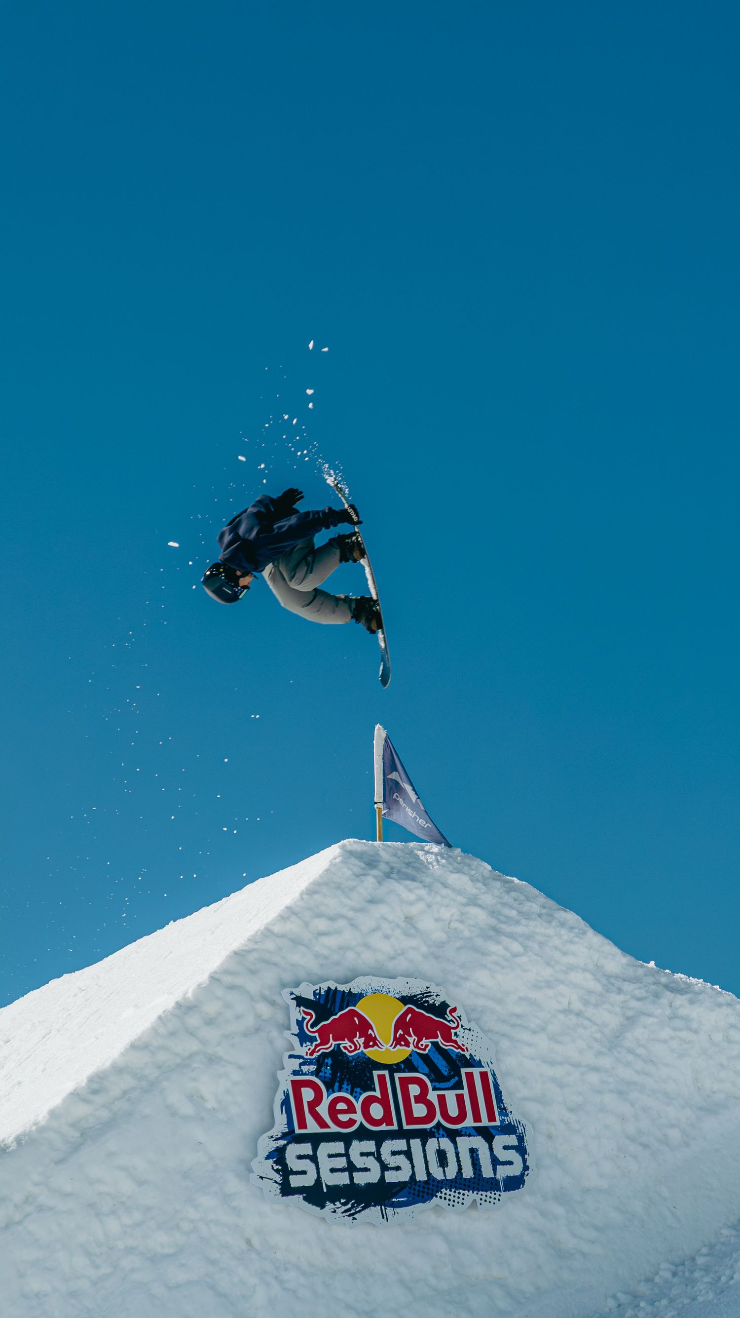 Snowboarder performs aerial trick over Red Bull Sessions branded snow ramp against clear blue sky.