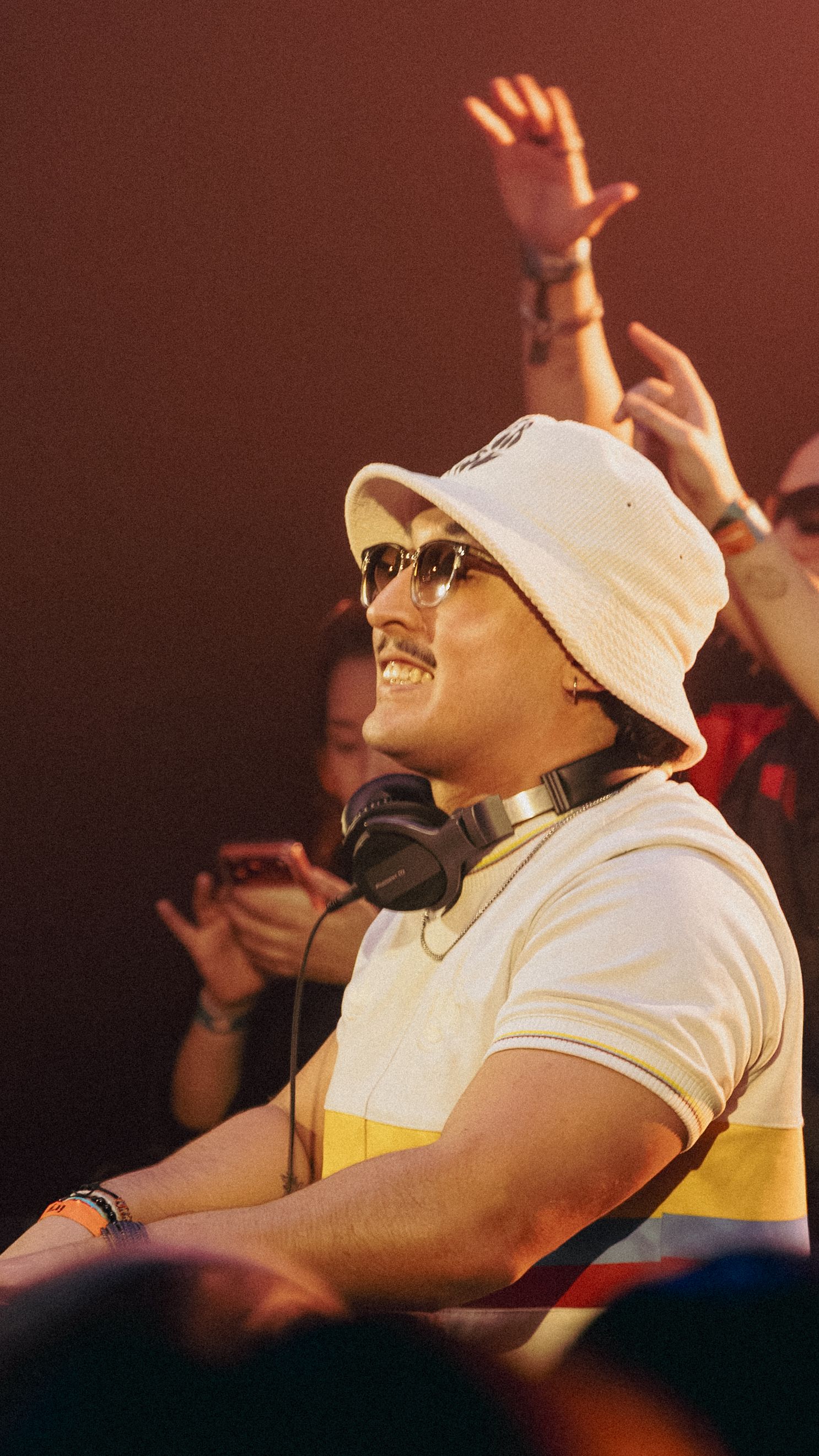 A smiling DJ wearing a white bucket hat, sunglasses, and headphones around his neck performs at a lively music event.