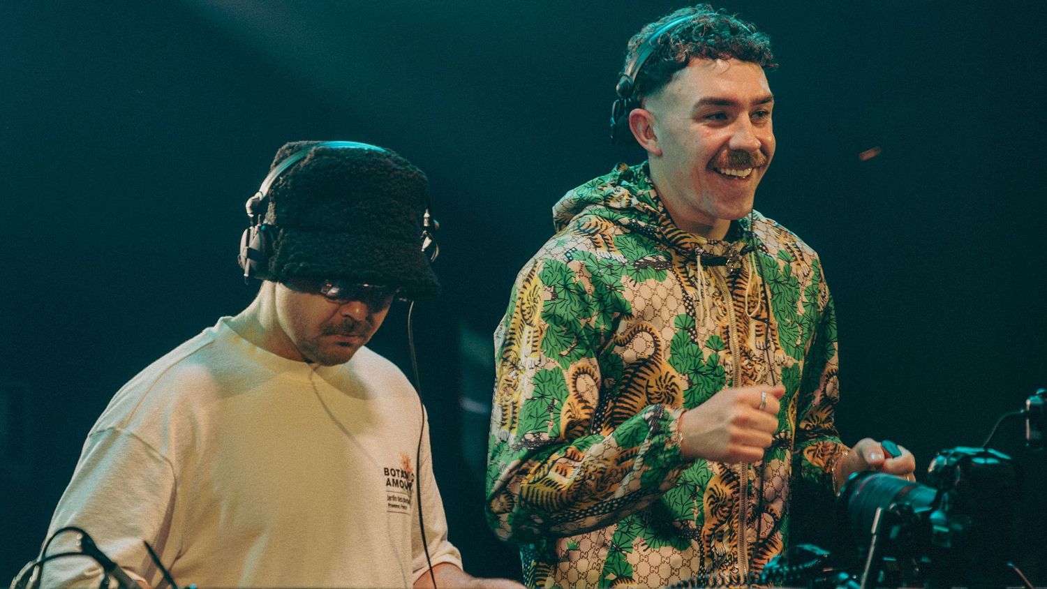 Two male DJs performing on stage, one wearing headphones, in colorful outfits under green stage lighting.