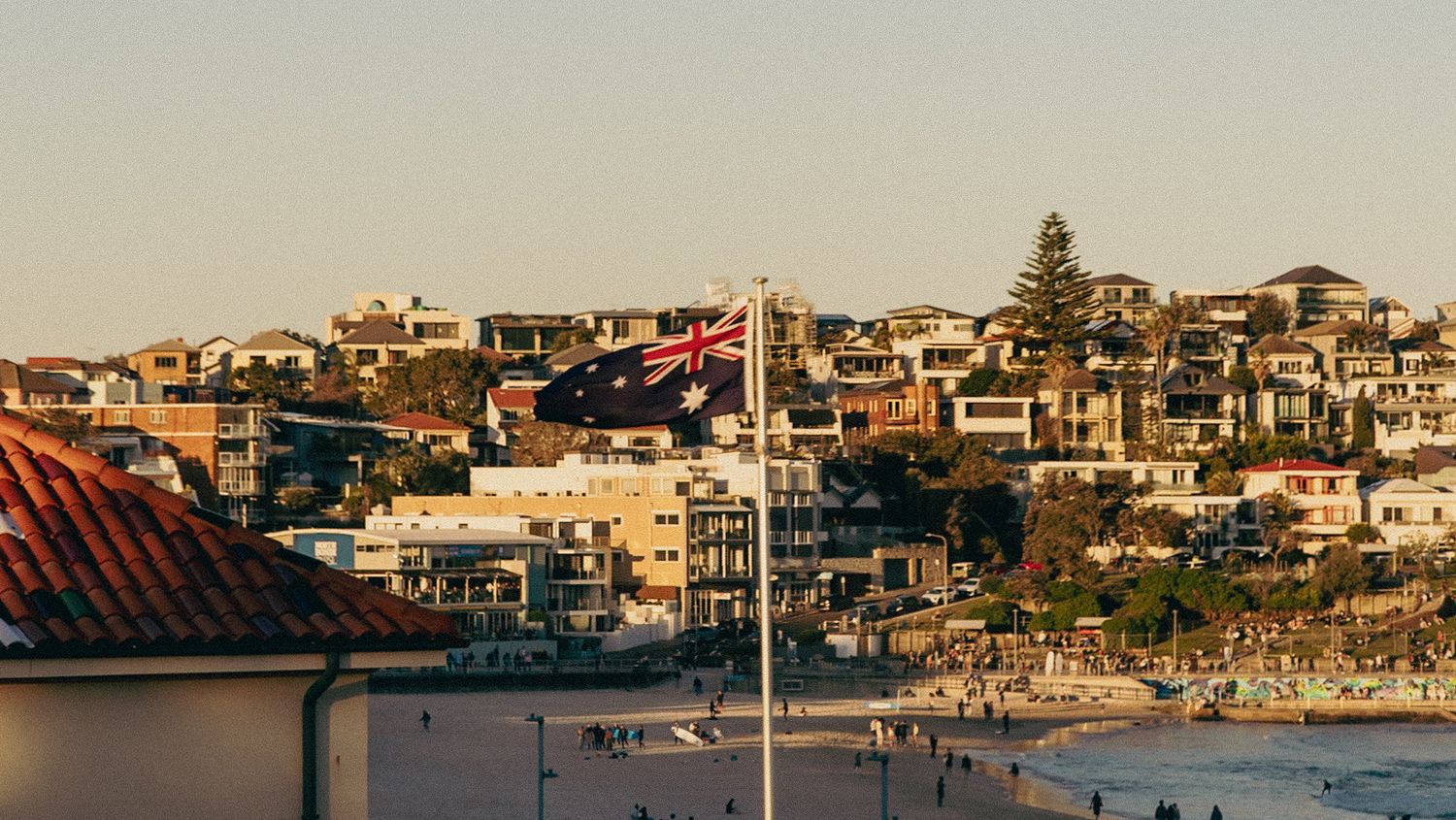 Australian flag flies over Bondi Beach with colorful hillside homes and golden sunset light in Sydney, Australia.