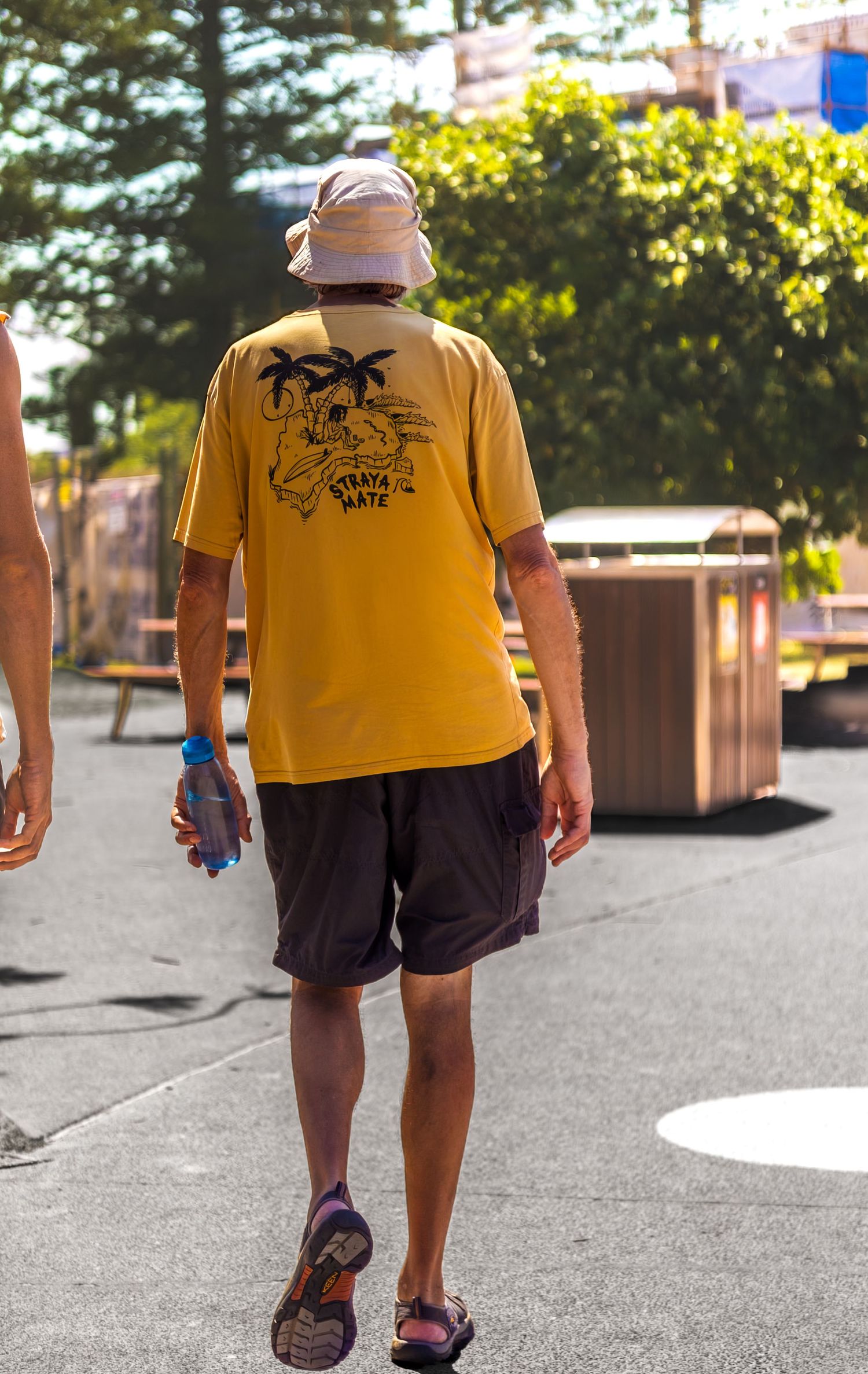 A person wearing a yellow t-shirt with palm tree design and brown shorts walking on a paved surface in daylight.