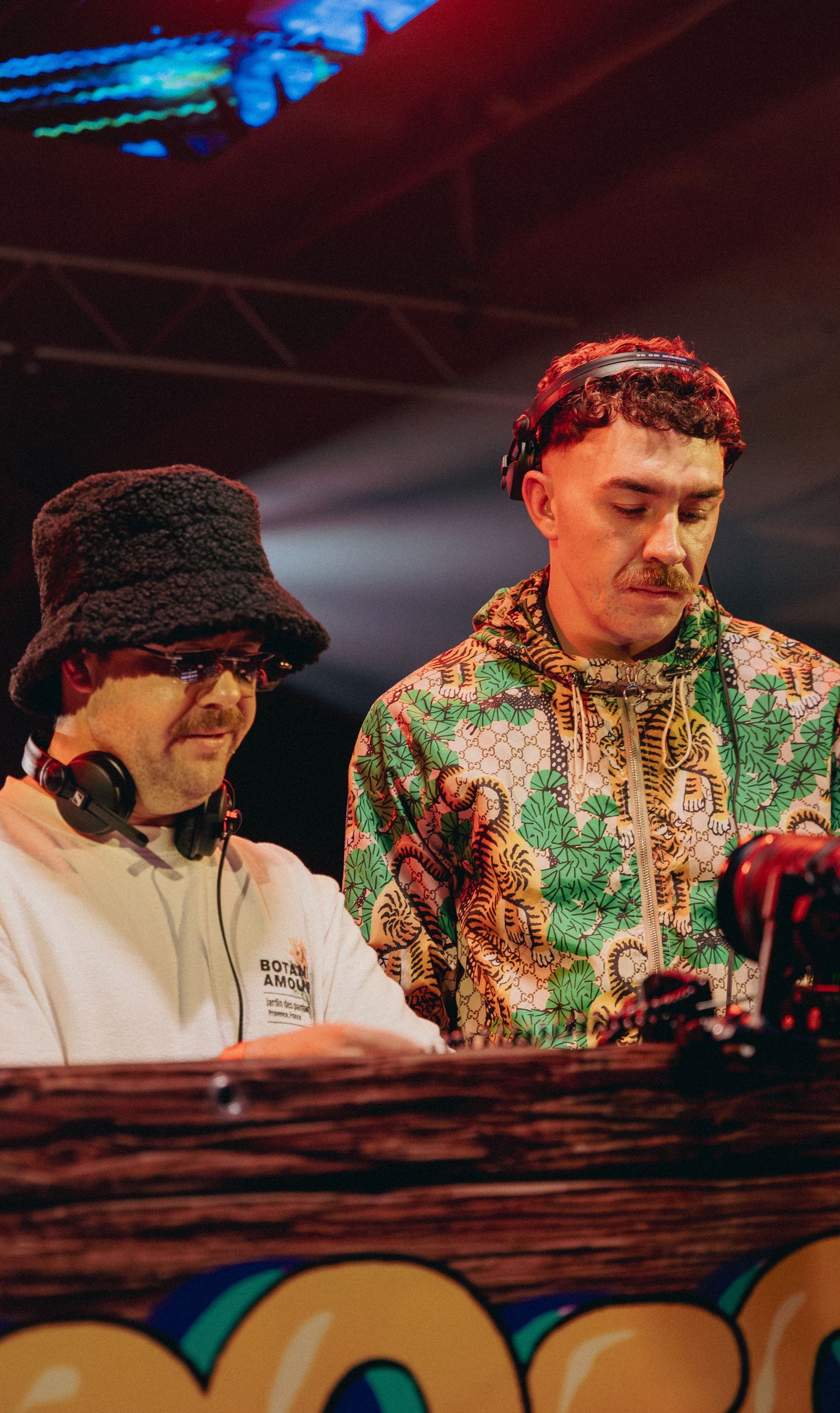 Two DJs performing at a festival, one wearing a bucket hat and headphones, the other in a colorful patterned hoodie.