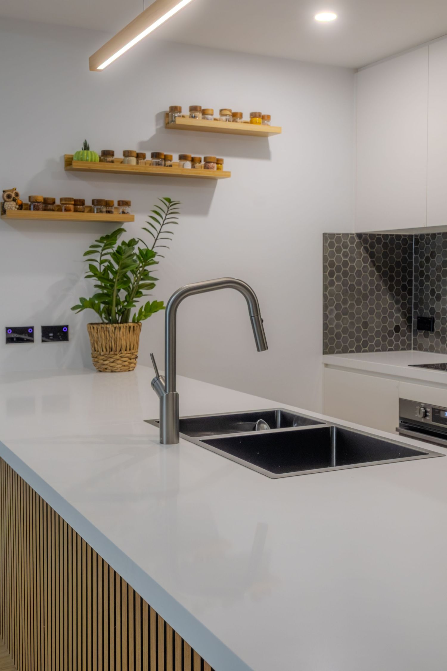 Modern white kitchen island with black sink, gooseneck faucet, wooden shelves with spice jars, and a potted green plant.