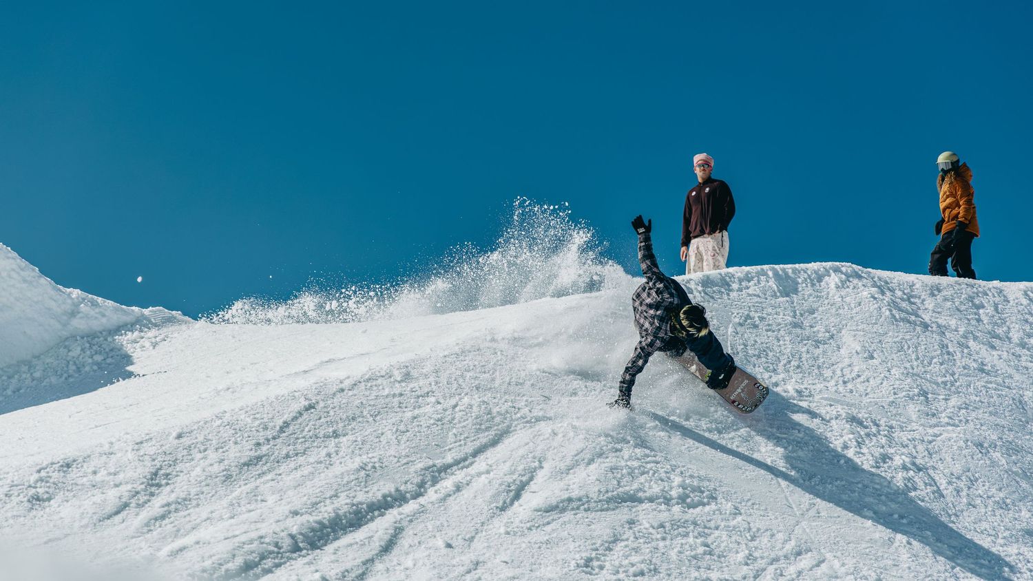 Snowboarder performing a trick on a snowy halfpipe with two spectators watching under a clear blue sky.