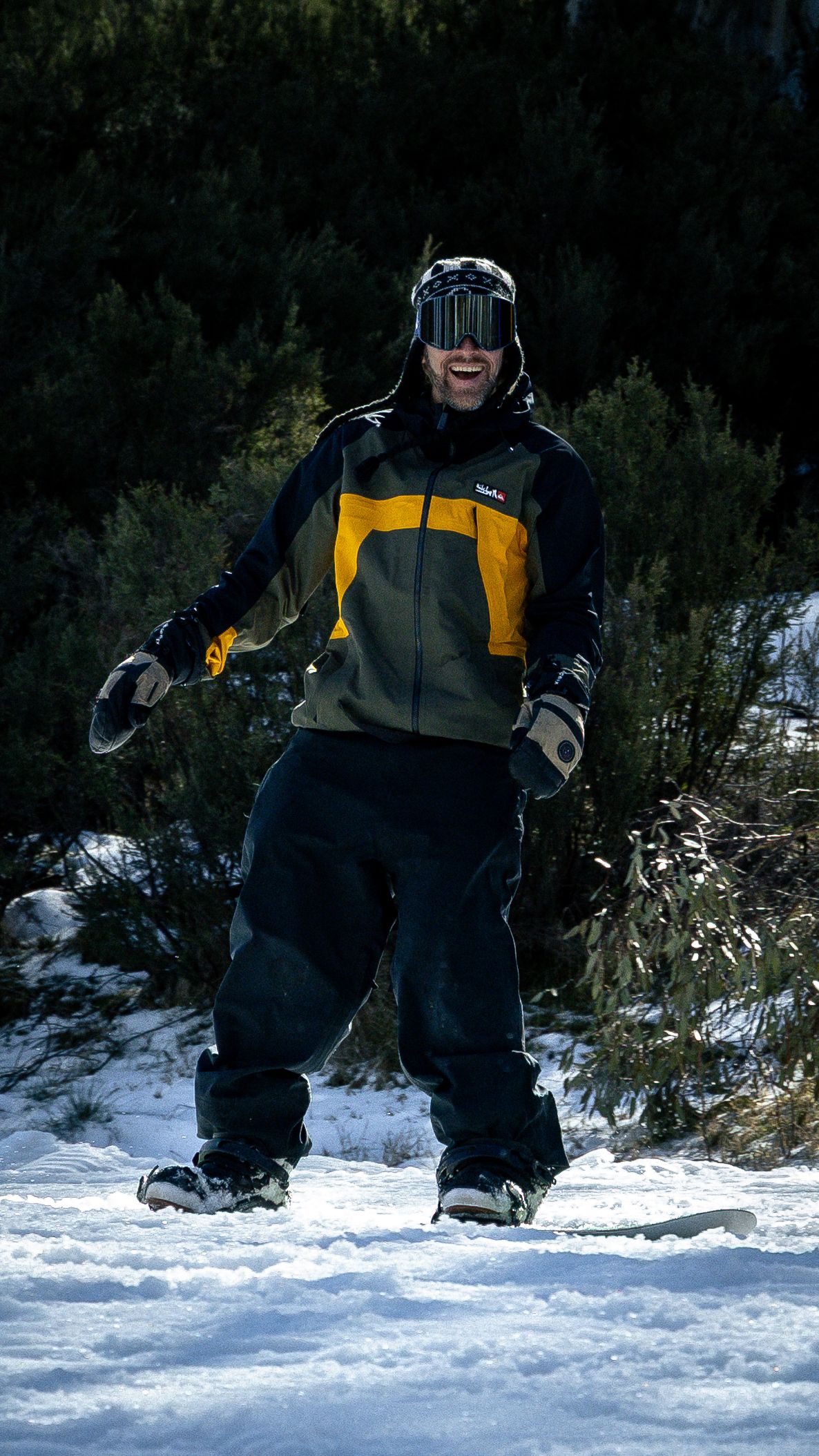 Smiling snowboarder in yellow and black jacket with goggles standing on snowy slope surrounded by evergreen trees.