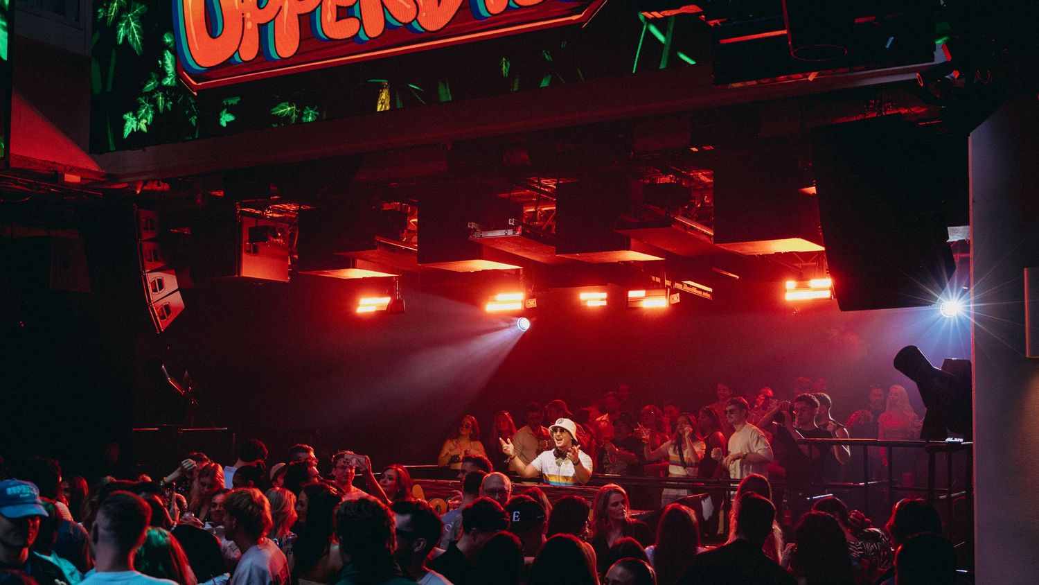 Crowded nightclub with DJ on stage, red neon lights, and energetic crowd dancing under colorful lighting.