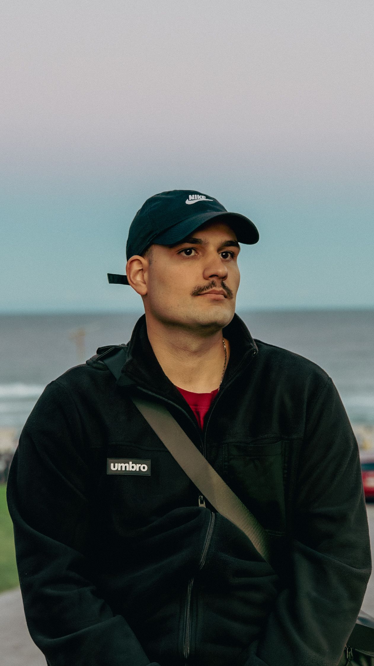 Young man with mustache wearing Nike cap and Umbro jacket standing near ocean at dusk.