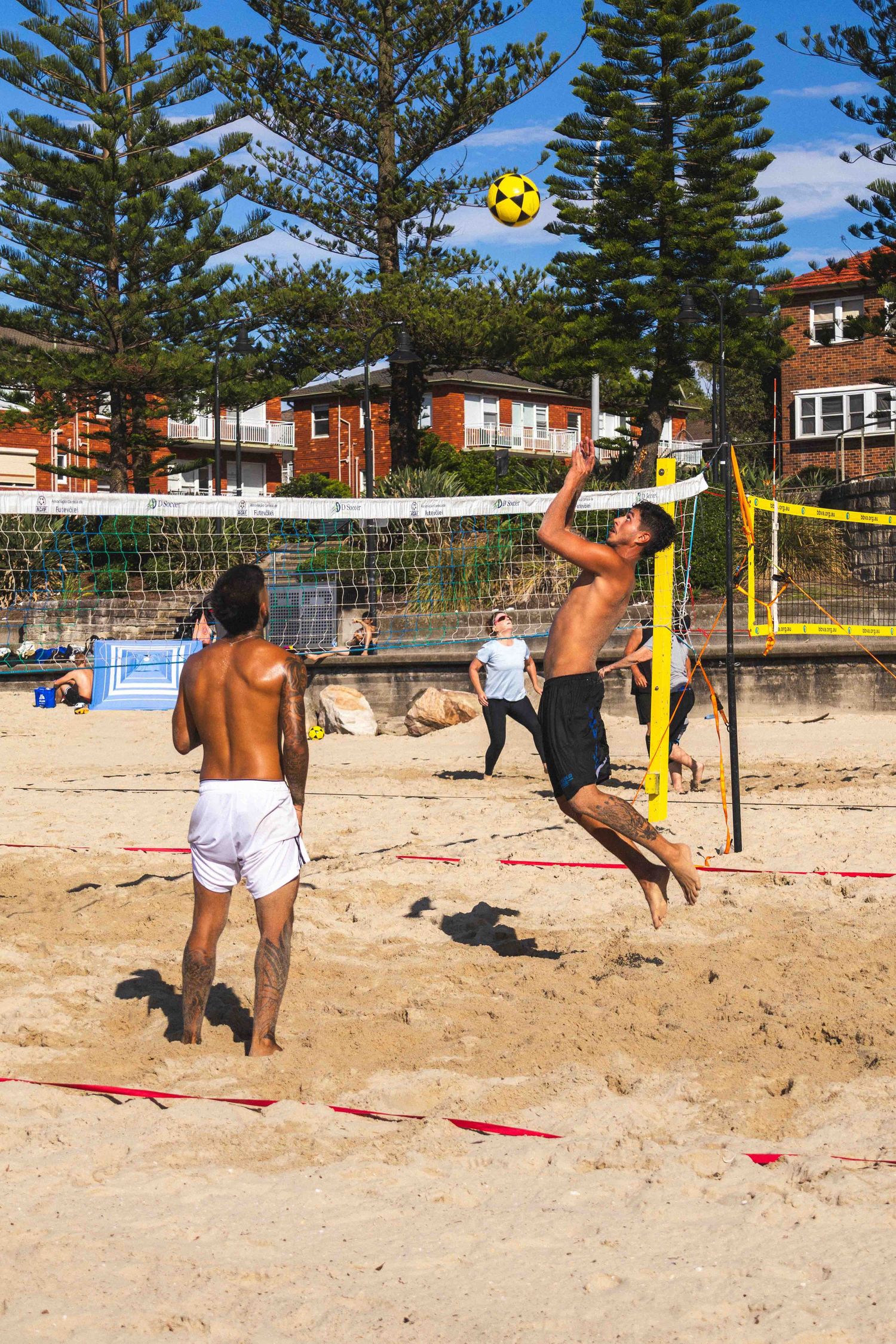 Beach volleyball players dive and jump for the ball during an intense match on a sandy court with palm trees in background.