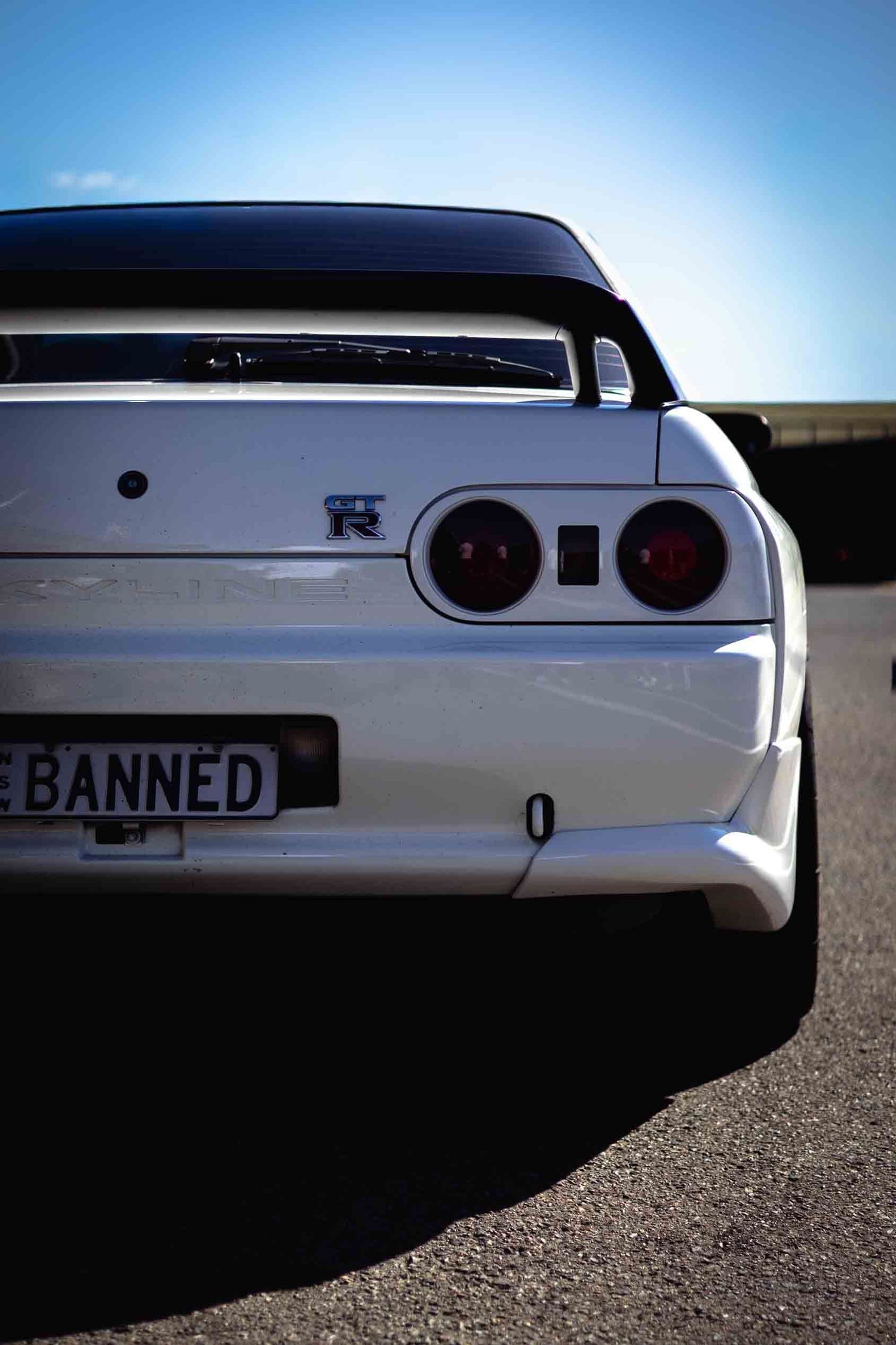 White Nissan Skyline GT-R R32 rear view with red tail lights and BANNED license plate on asphalt.