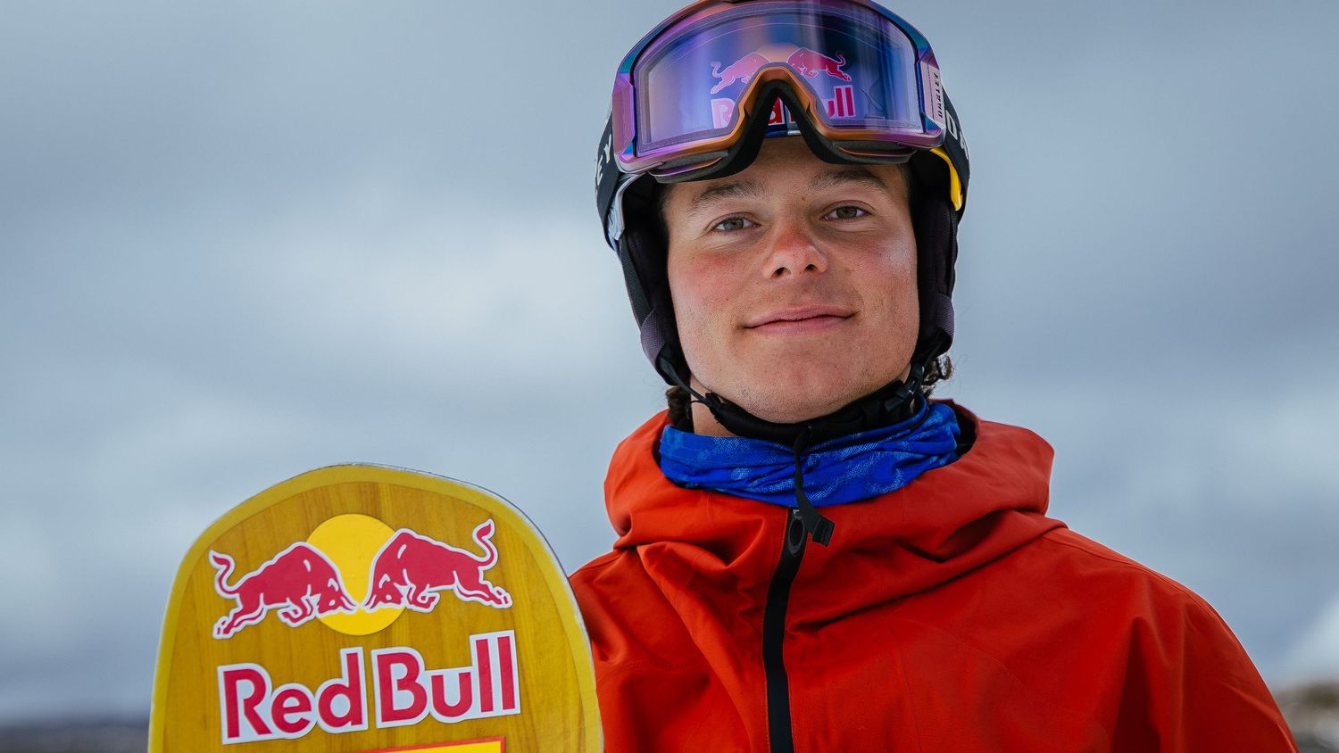 Young snowboarder in red jacket holds a yellow Red Bull snowboard, wearing a helmet and goggles outdoors.