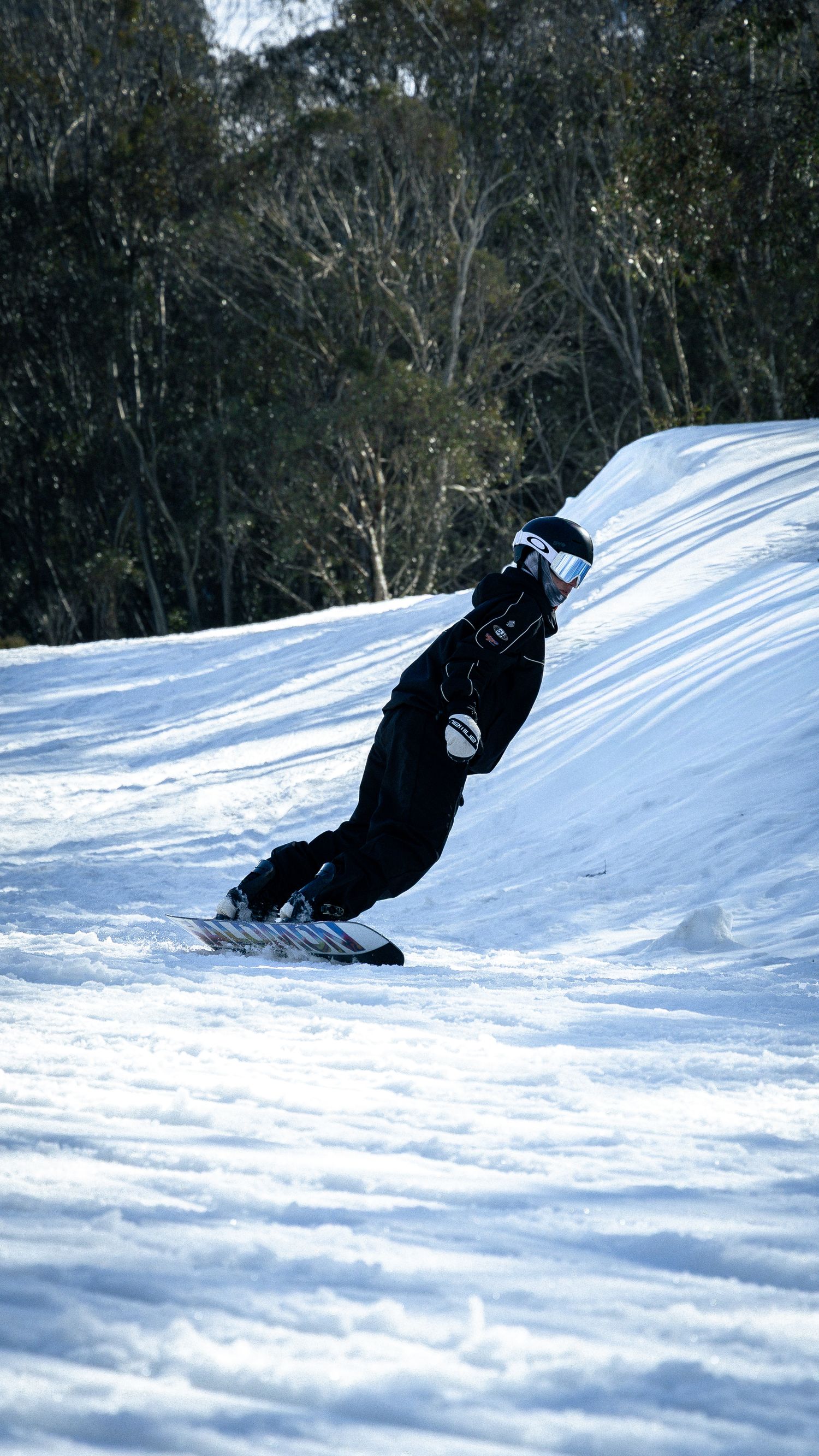 Snowboarder in black gear carving down a snowy slope with trees in the background on a sunny winter day.