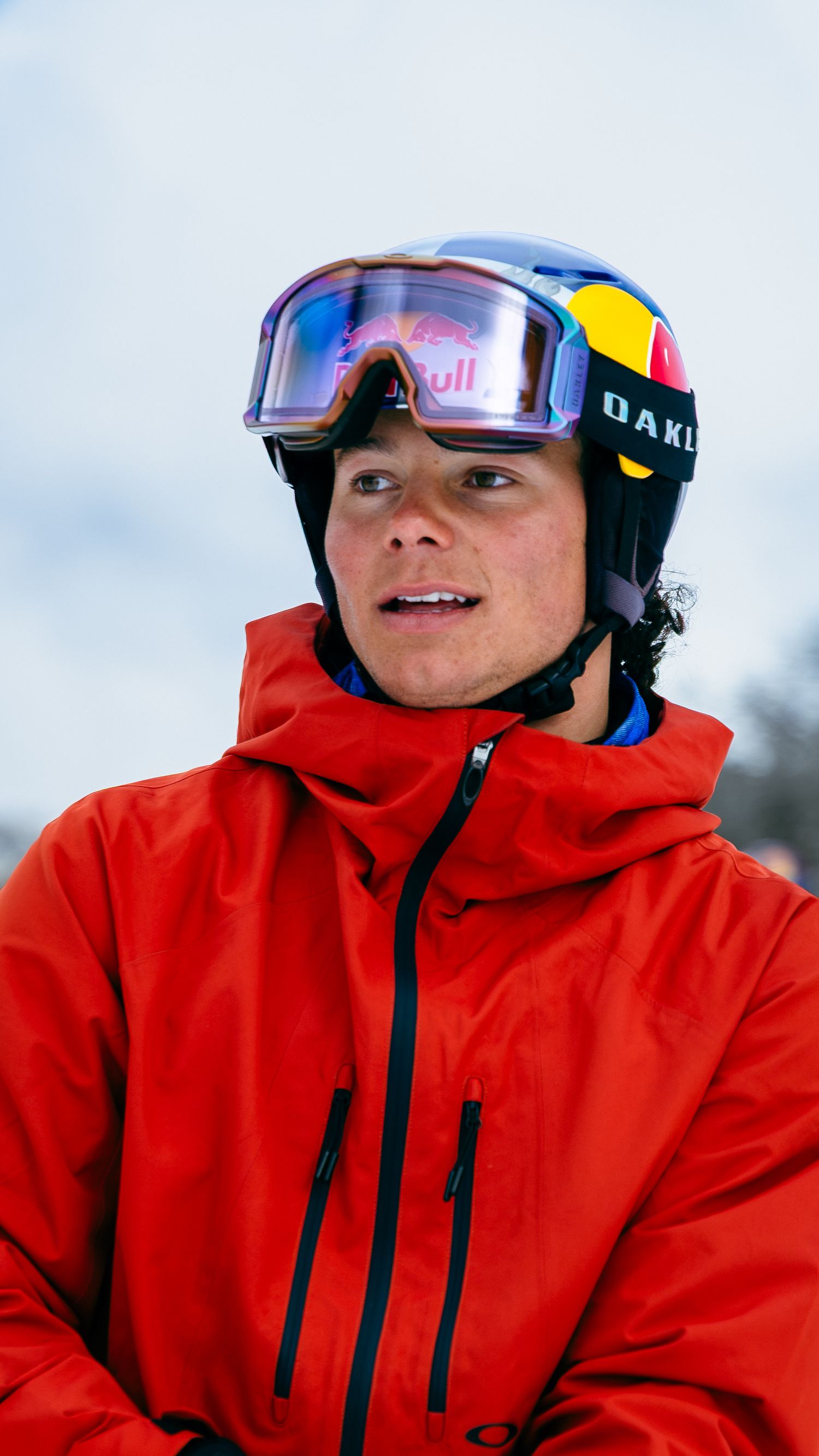 Young male skier wearing a red jacket, Oakley goggles, and a Red Bull helmet on a snowy slope.