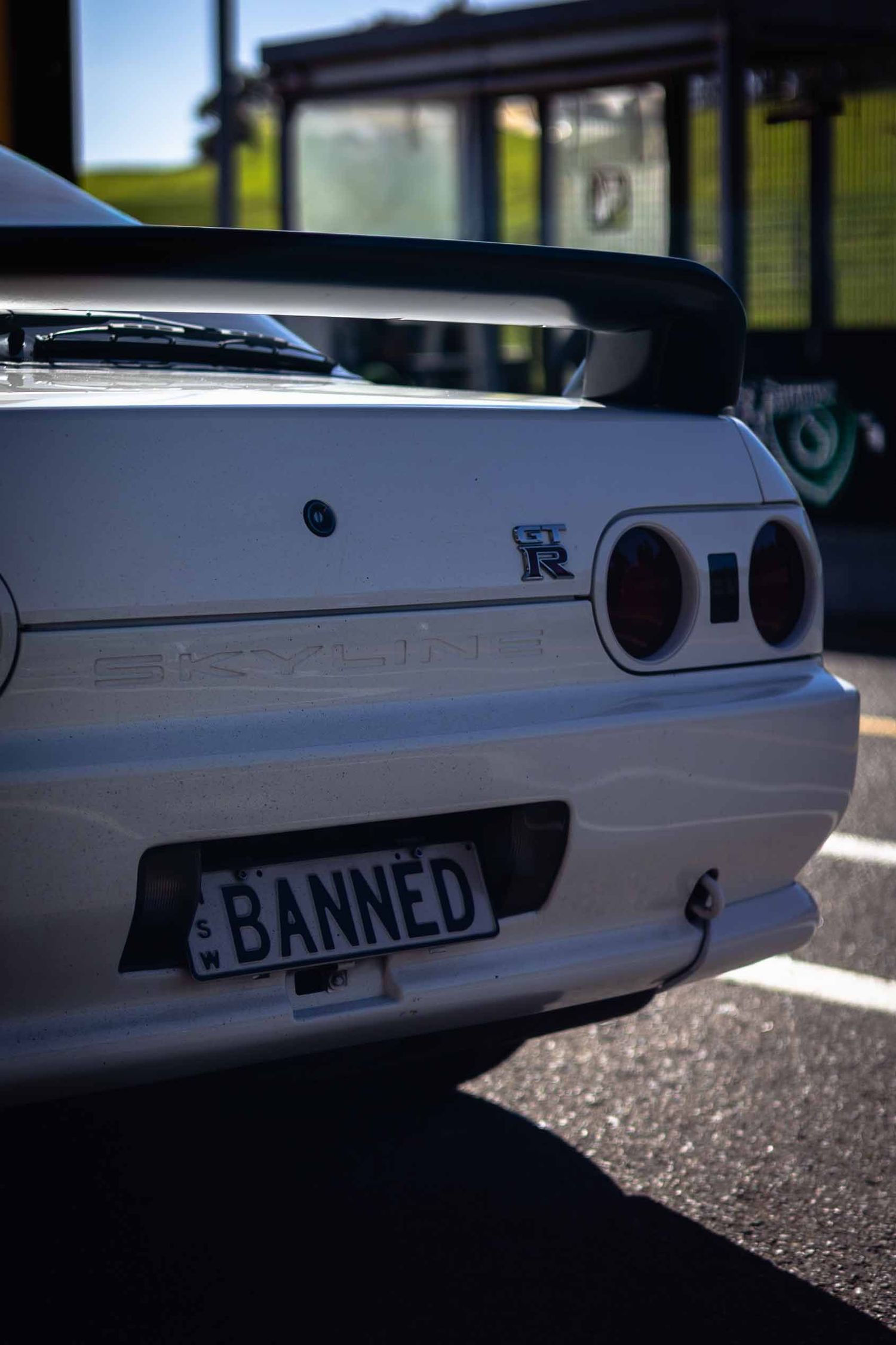 White Nissan Skyline GT-R R32 rear view with BANNED custom license plate parked in sunlight.