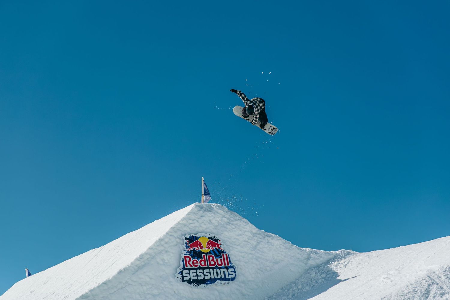 Snowboarder performs high aerial trick above a Red Bull Sessions branded snow ramp against a clear blue sky.