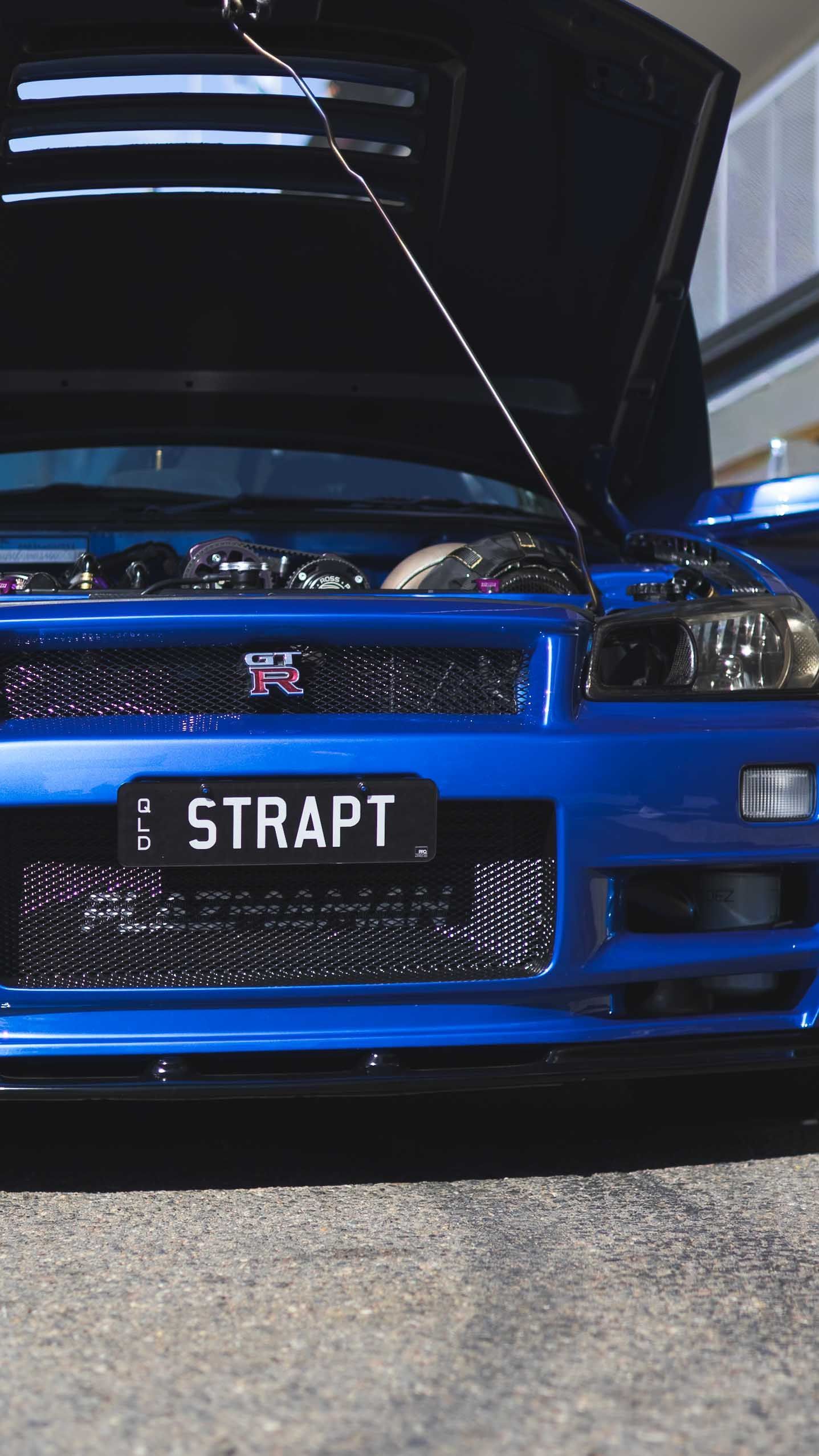 Blue Nissan Skyline GT-R R34 with hood open, displaying engine, custom plate STRAPT, at a car show.