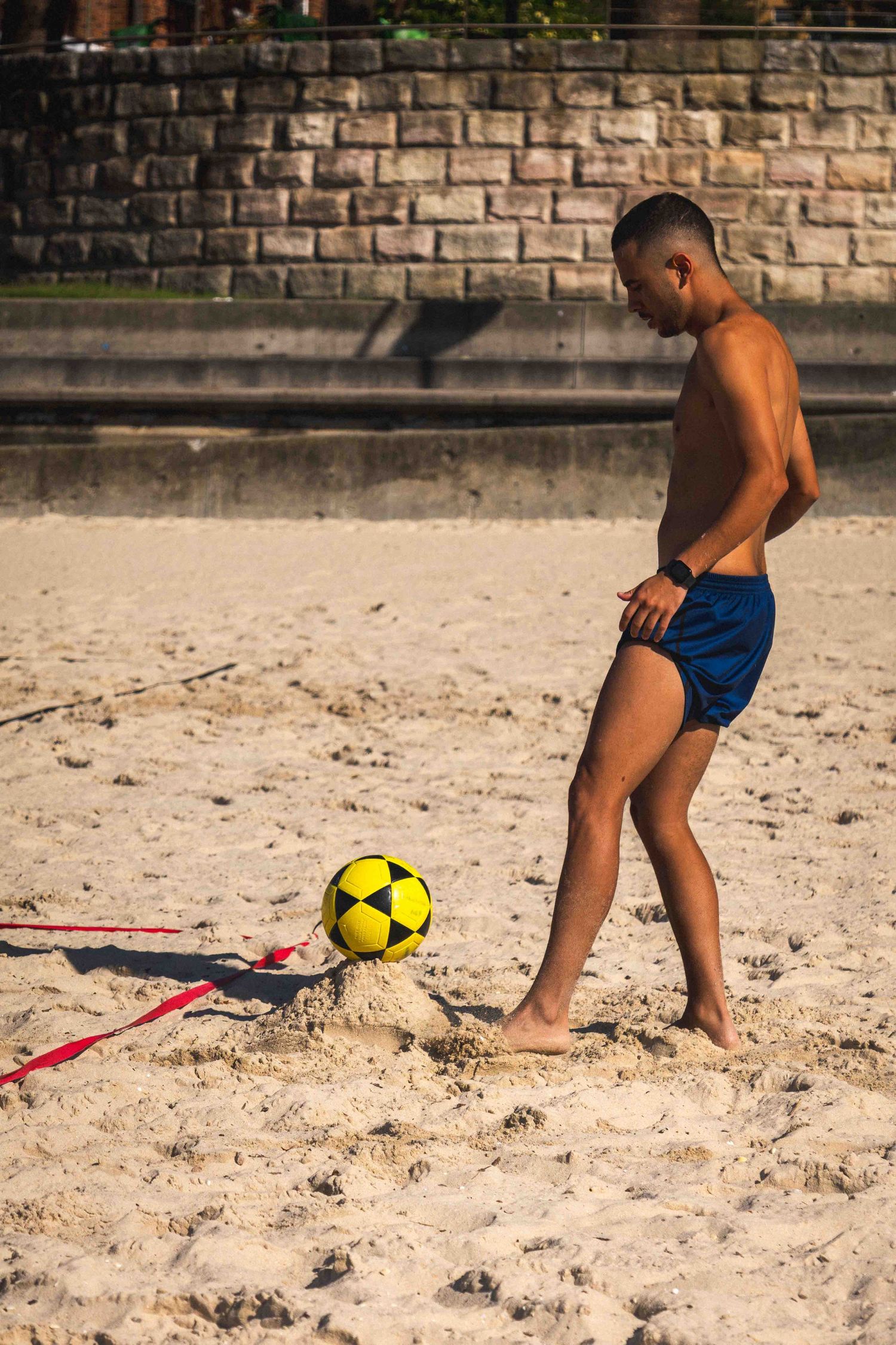 A young child in blue shorts plays with a yellow soccer ball on a sandy beach near a stone wall.