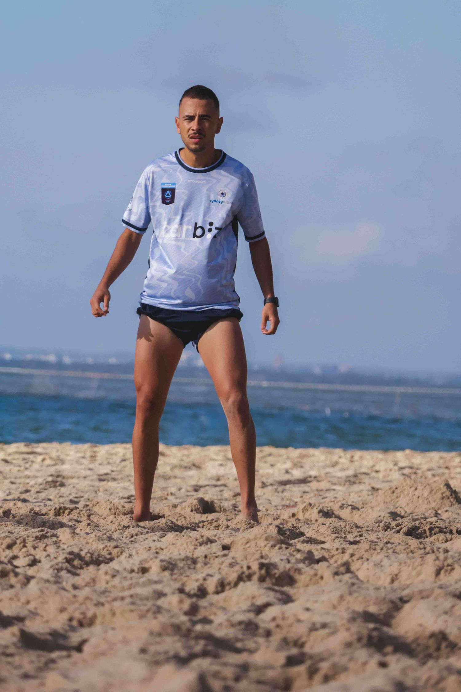Man in white jersey and black swim briefs standing on sandy beach with ocean in background.