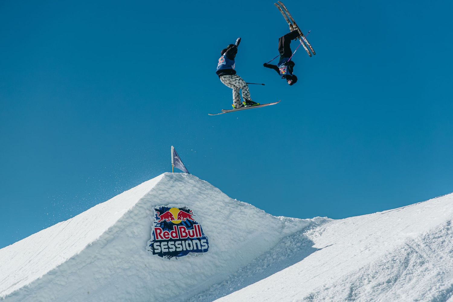 Two snowboarders perform aerial tricks over a snow jump at the Red Bull Sessions competition under a clear blue sky.