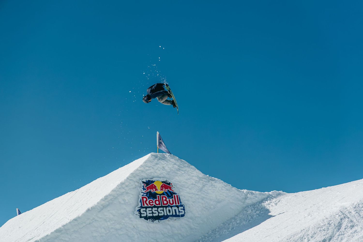 Snowboarder performs a high aerial trick over a Red Bull Sessions branded snow jump against a clear blue sky.