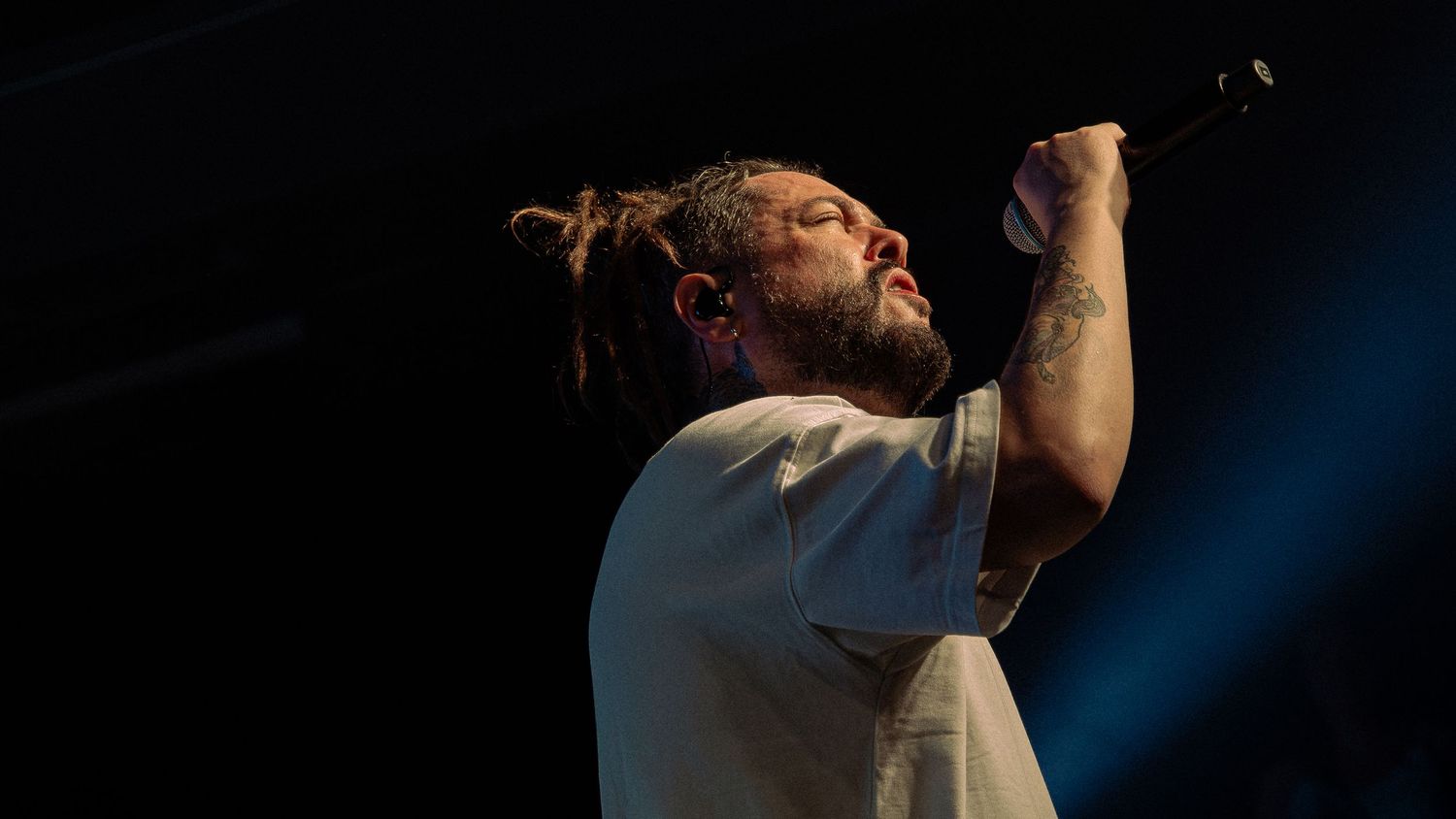 Tattooed male singer with dreadlocks performing on stage, holding microphone up under blue stage lights.