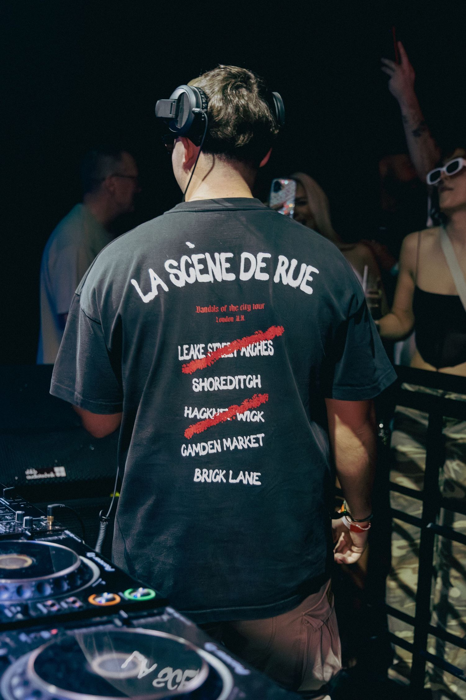 DJ wearing La Scène De Rue graphic tee at mixing decks in dark nightclub setting.