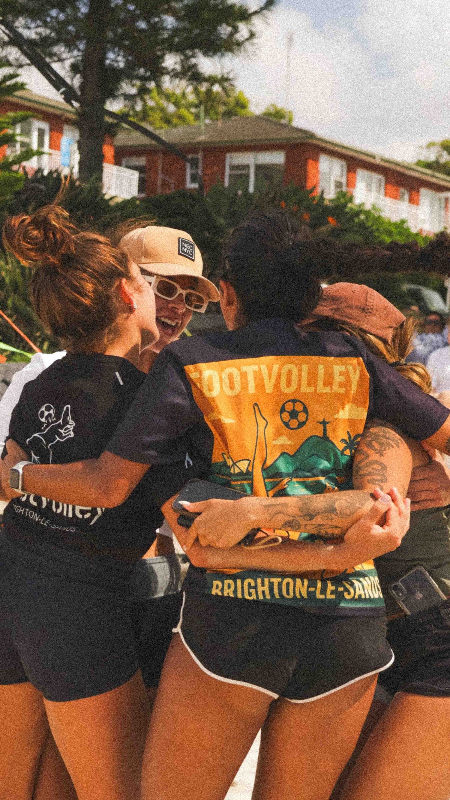 Group of friends in DotVolley Brighton-le-Sands jerseys celebrating in a team huddle on a sunny beach day.