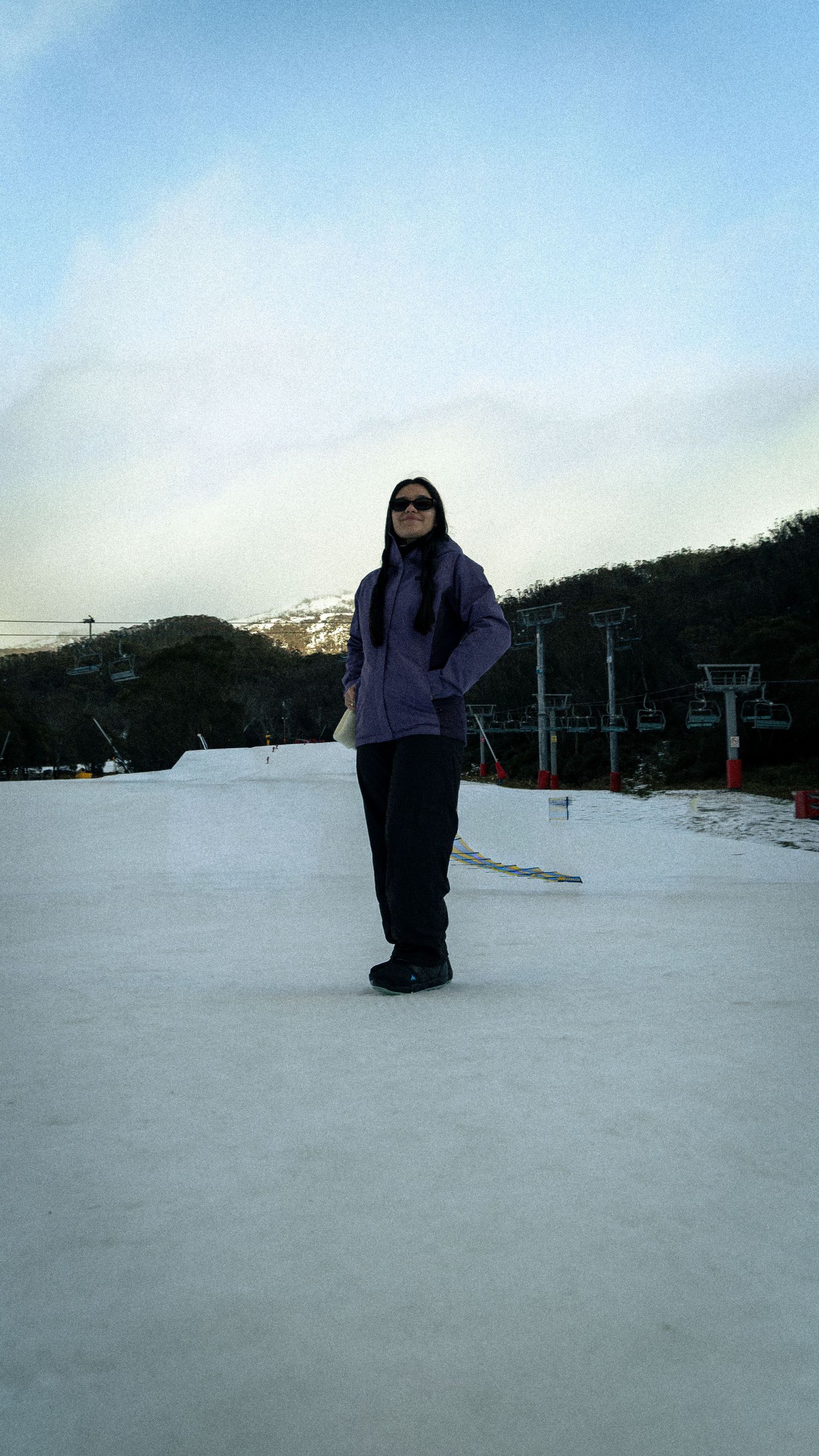 Woman in purple jacket and sunglasses standing on snowy ski slope with mountain and ski lift in background.
