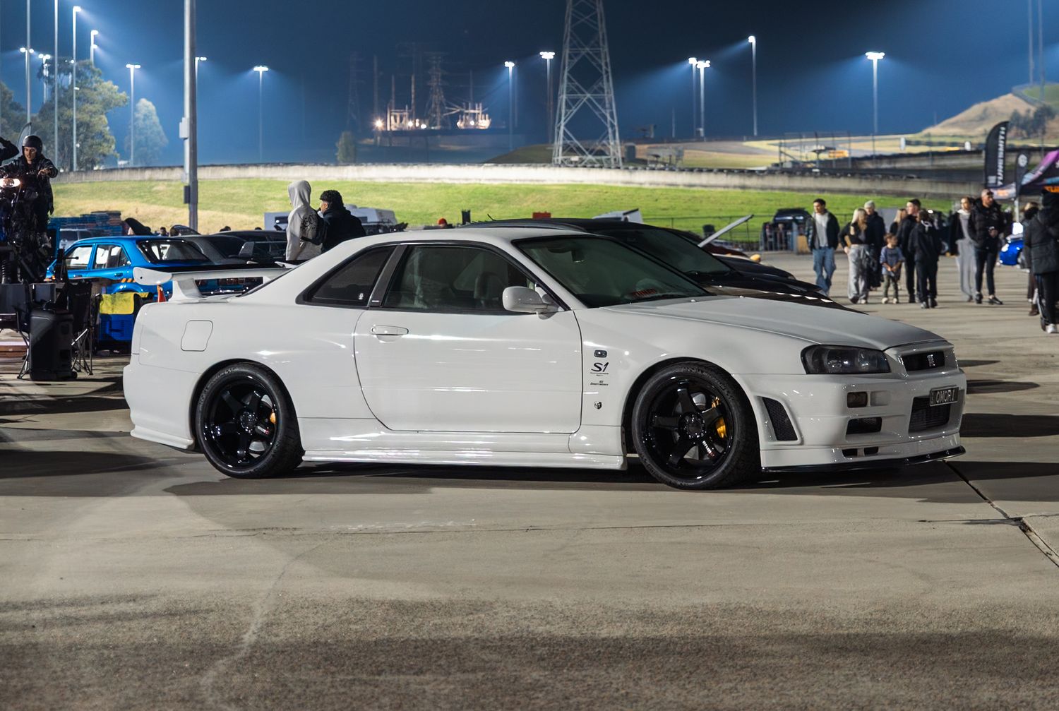White Nissan Skyline R34 GT-R sports car parked at a night racing event on a dark track.