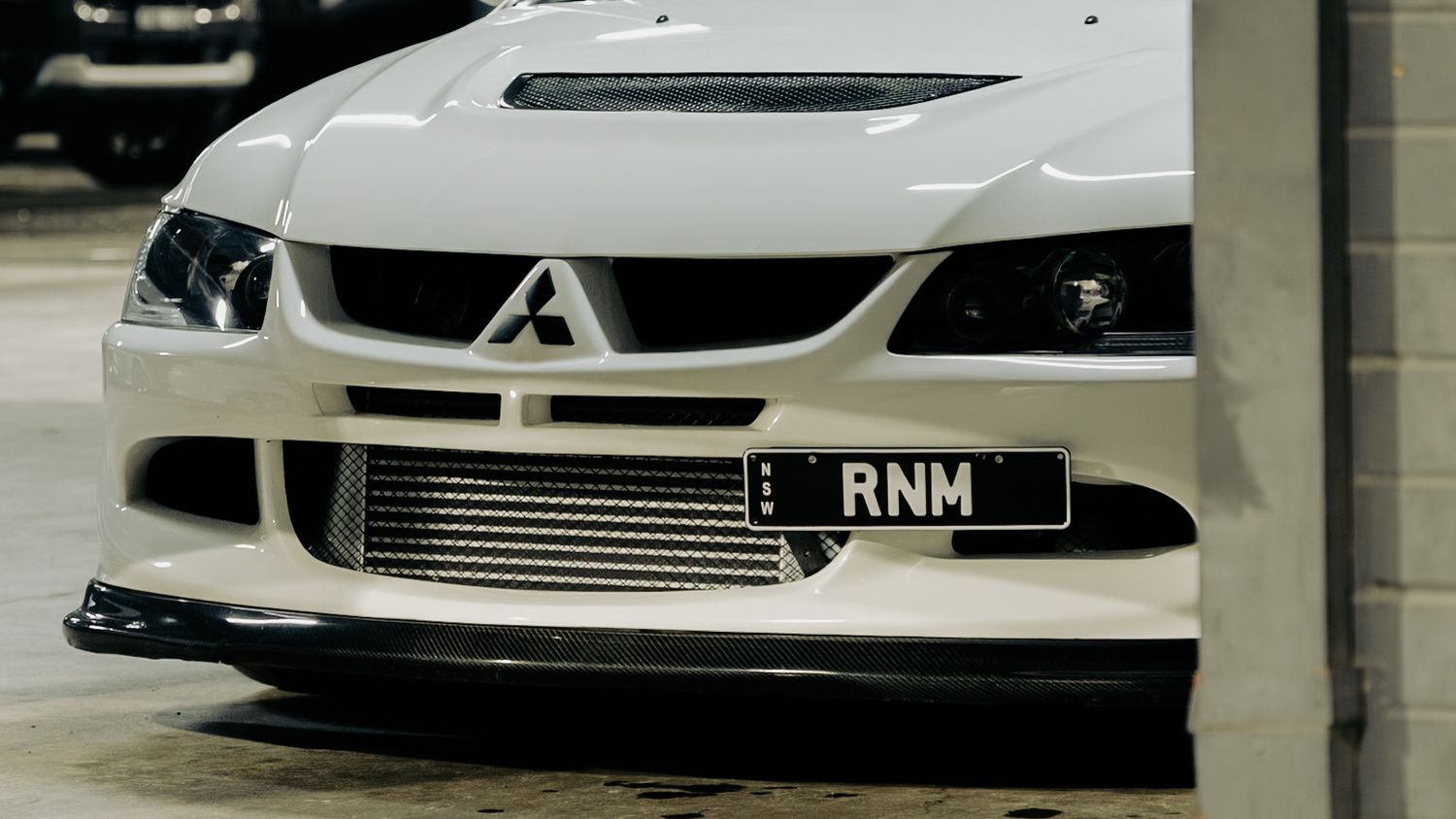 White Mitsubishi Lancer Evolution front bumper with carbon fiber lip spoiler and RNM license plate in a parking garage.