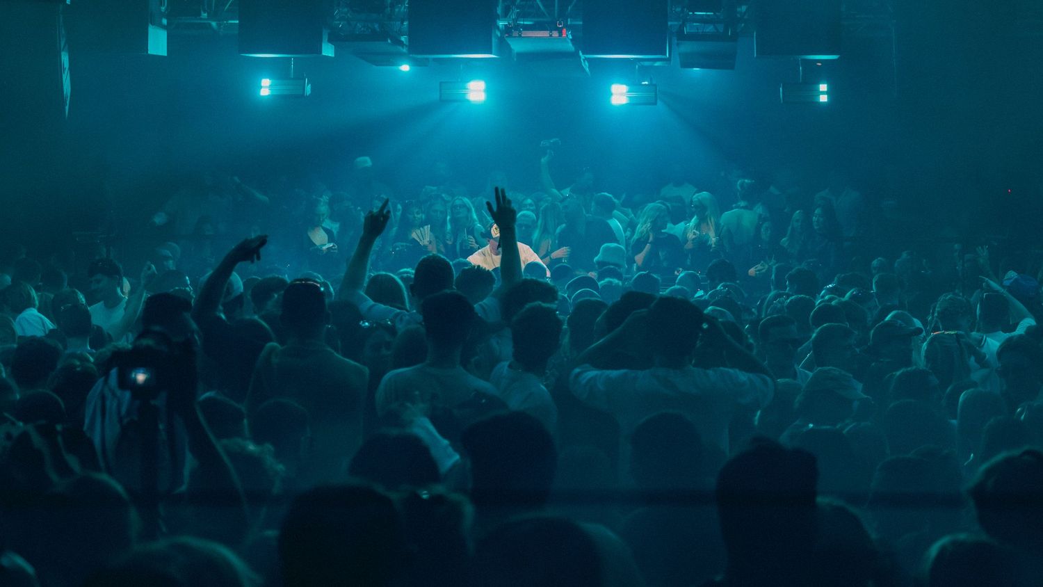 Crowd of concert-goers with raised hands under blue stage lights in a packed nightclub venue.